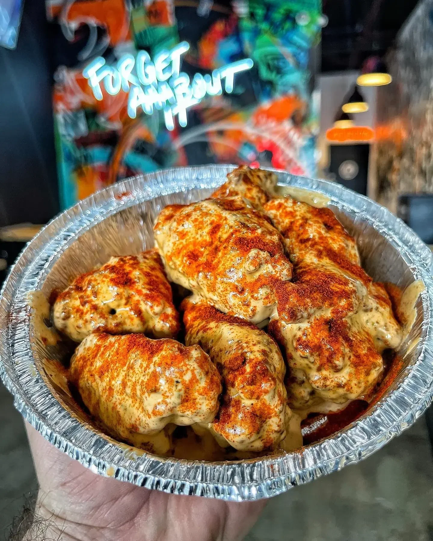 A hand holding a round aluminum container filled with chicken wings sprinkled with red seasoning, with a background featuring colorful graffiti art and the words 'Forget About It' in bright blue neon-style lettering.