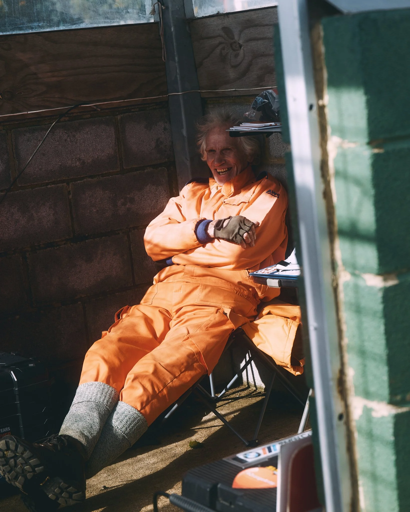 An elderly person in an orange jumpsuit sitting on a folding chair, smiling and leaning back with arms crossed, in a rustic outdoor setting.