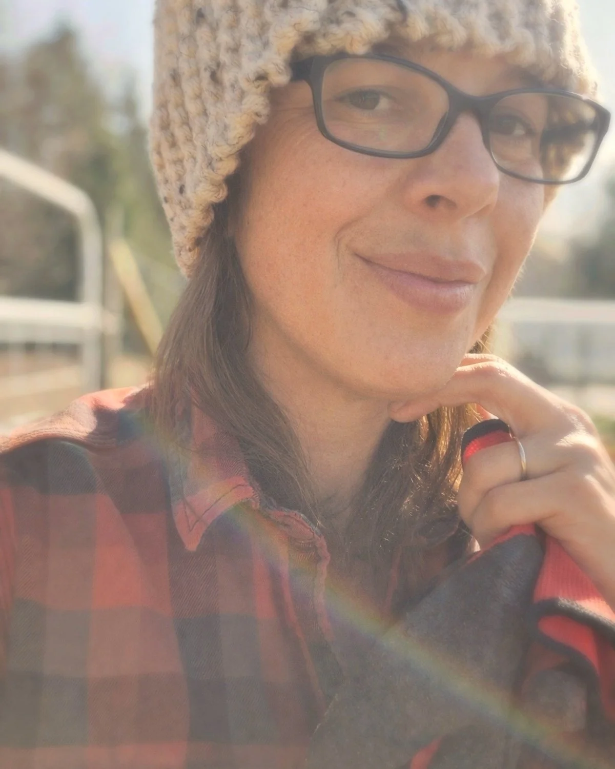 Close-up of a woman wearing glasses and a knit hat, outdoors in sunlight, smiling gently with a rainbow-like flare.