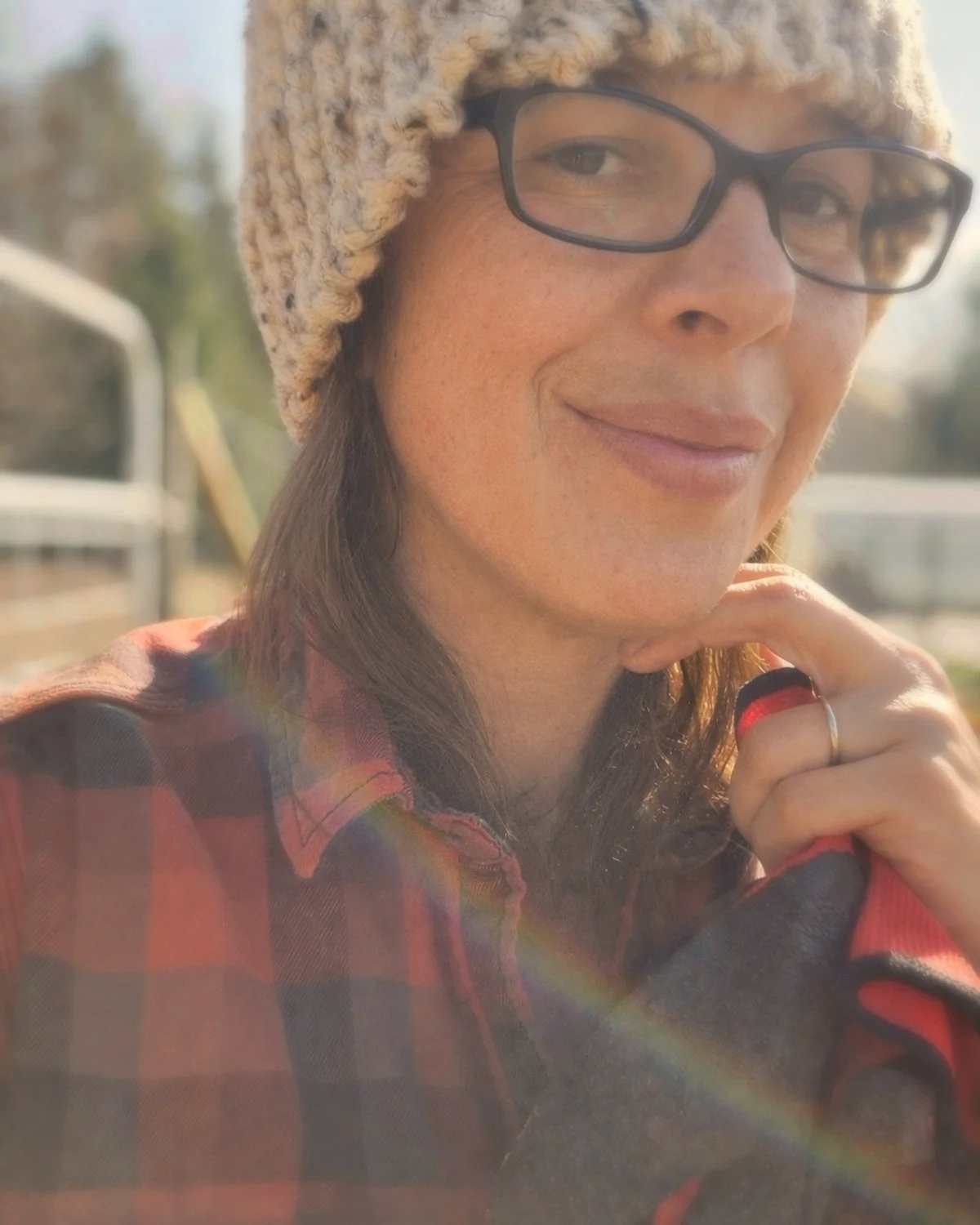 A woman with glasses and a knit hat smiling outdoors, with sunlight creating a rainbow lens flare in front of her.