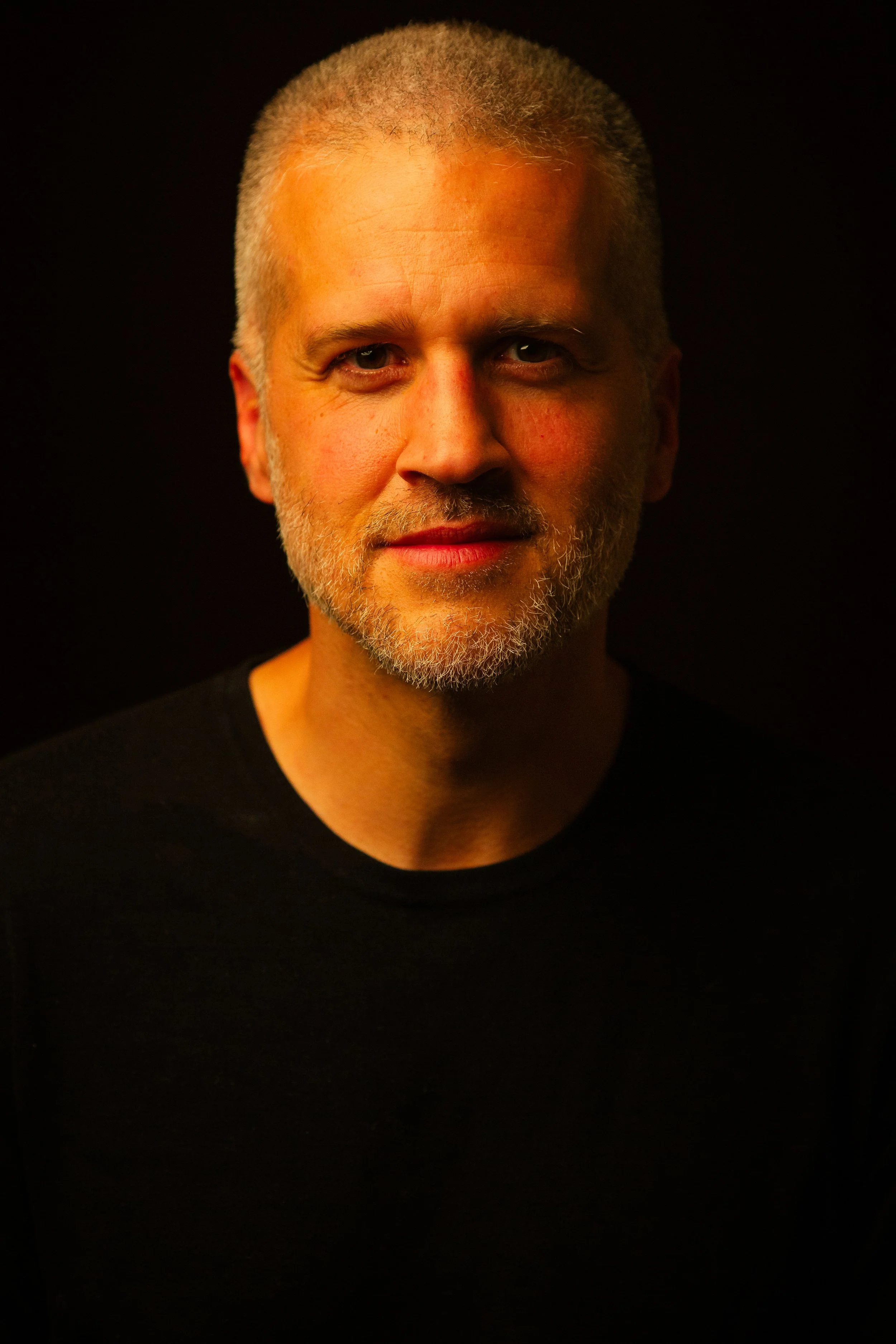 A middle-aged man with a closely shaved head, gray beard, and light skin, wearing a black shirt, looking into the camera against a dark background.