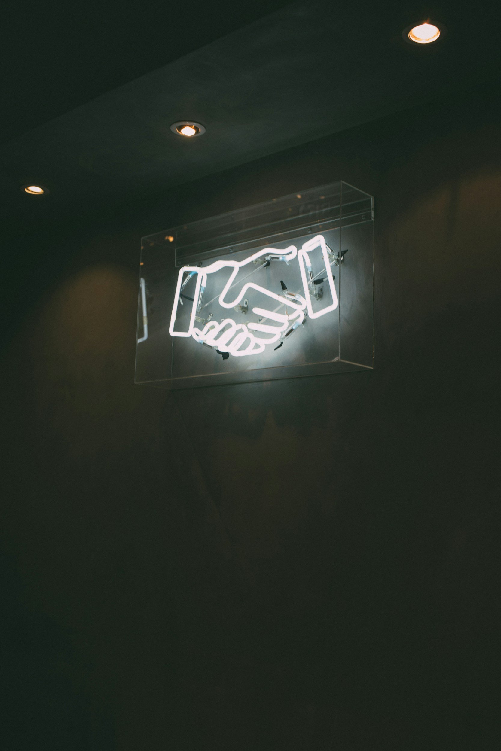 A neon sign of two hands shaking, mounted on a transparent box against a dark wall.