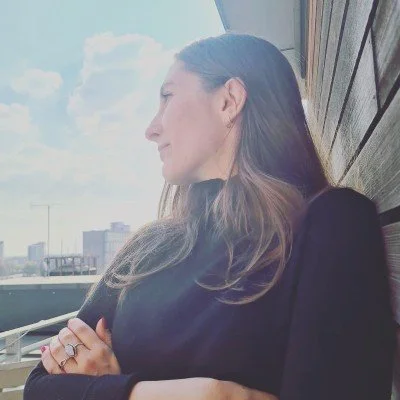 Side profile of a woman with long brown hair, sitting outdoors on a balcony, looking at the sky with a city skyline in the background.