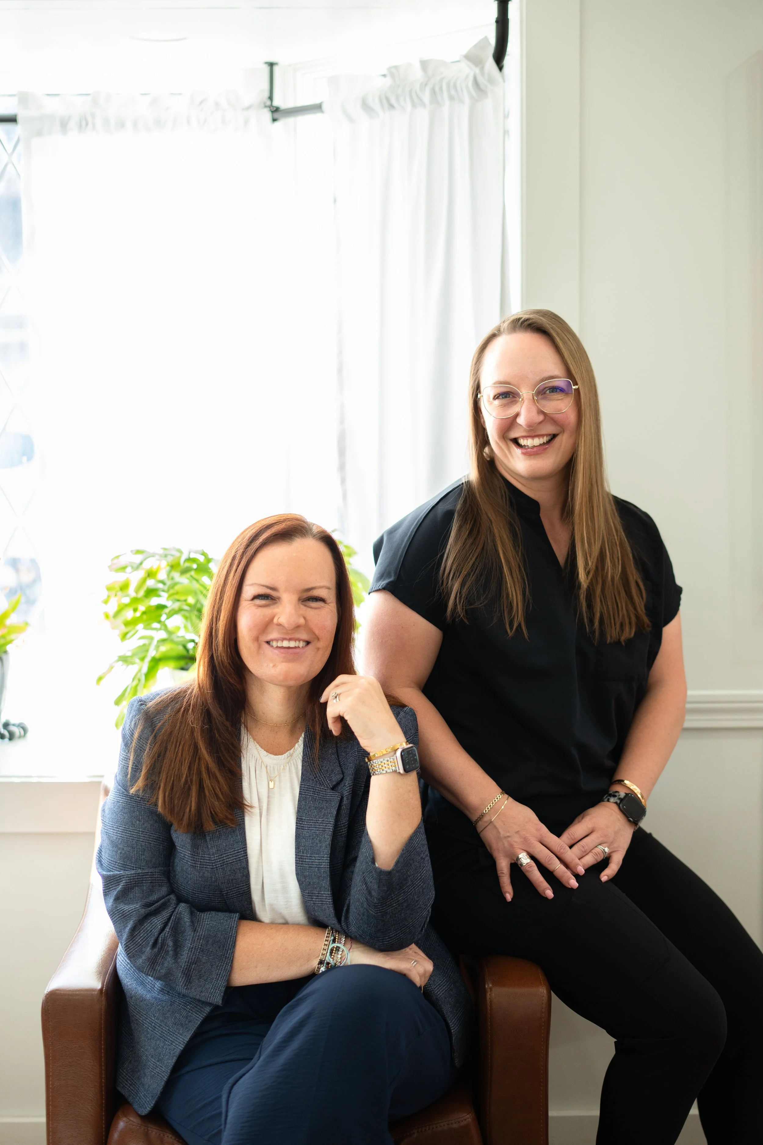 Two women smiling in a well-lit room with a window and plants in the background.