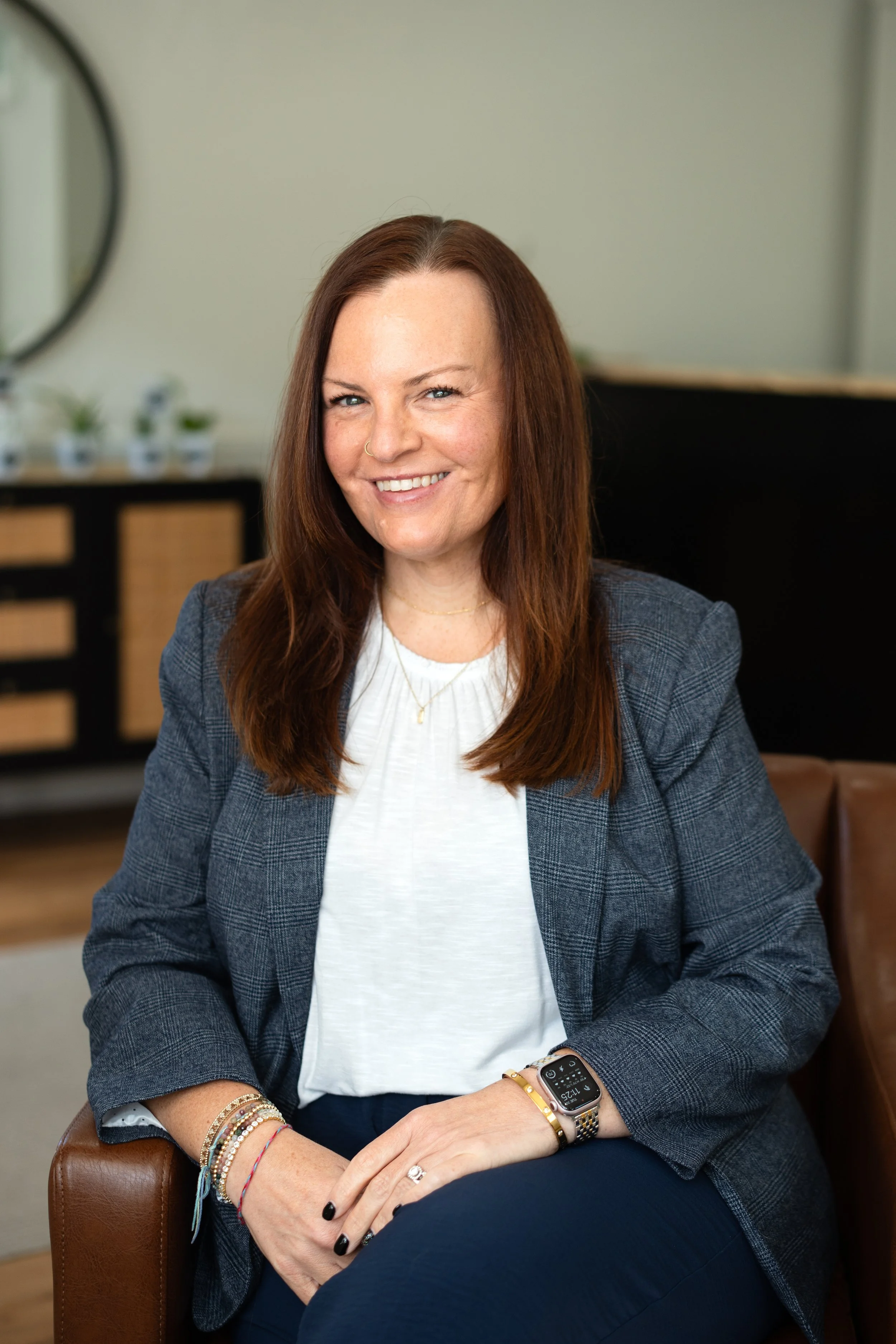 A woman with long brown hair, smiling, sitting on a brown leather chair in an office or lounge with a neutral-colored wall, a mirror, and some small potted plants in the background.
