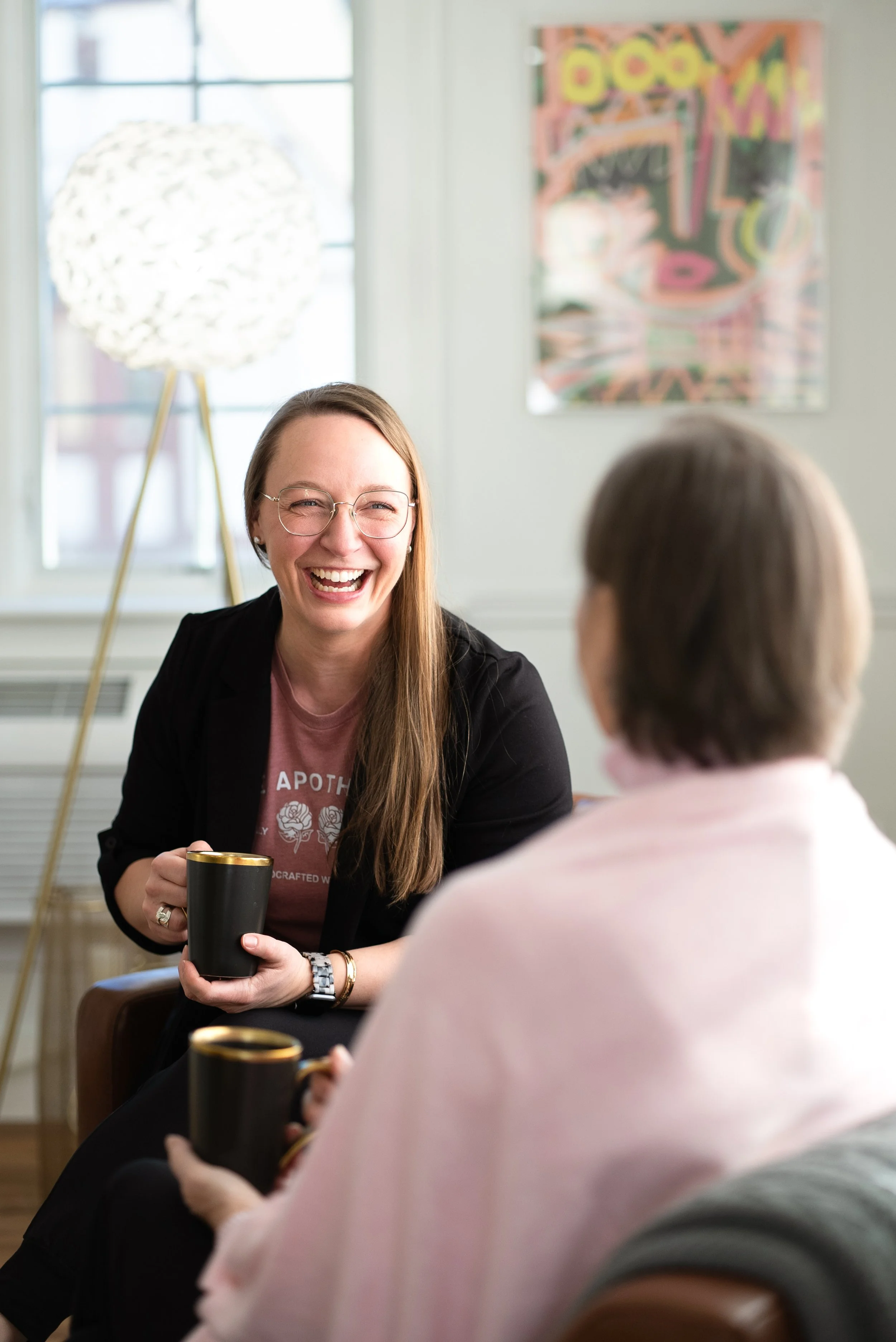 Two women having a friendly conversation in a cozy, bright room. One woman with long red hair, glasses, a pink T-shirt, and a black jacket smiling and holding a black mug. The other woman, seen from behind, has short hair and wears a light pink shawl, also holding a black mug. There is a window, a decorative lamp, and an abstract colorful poster in the background.