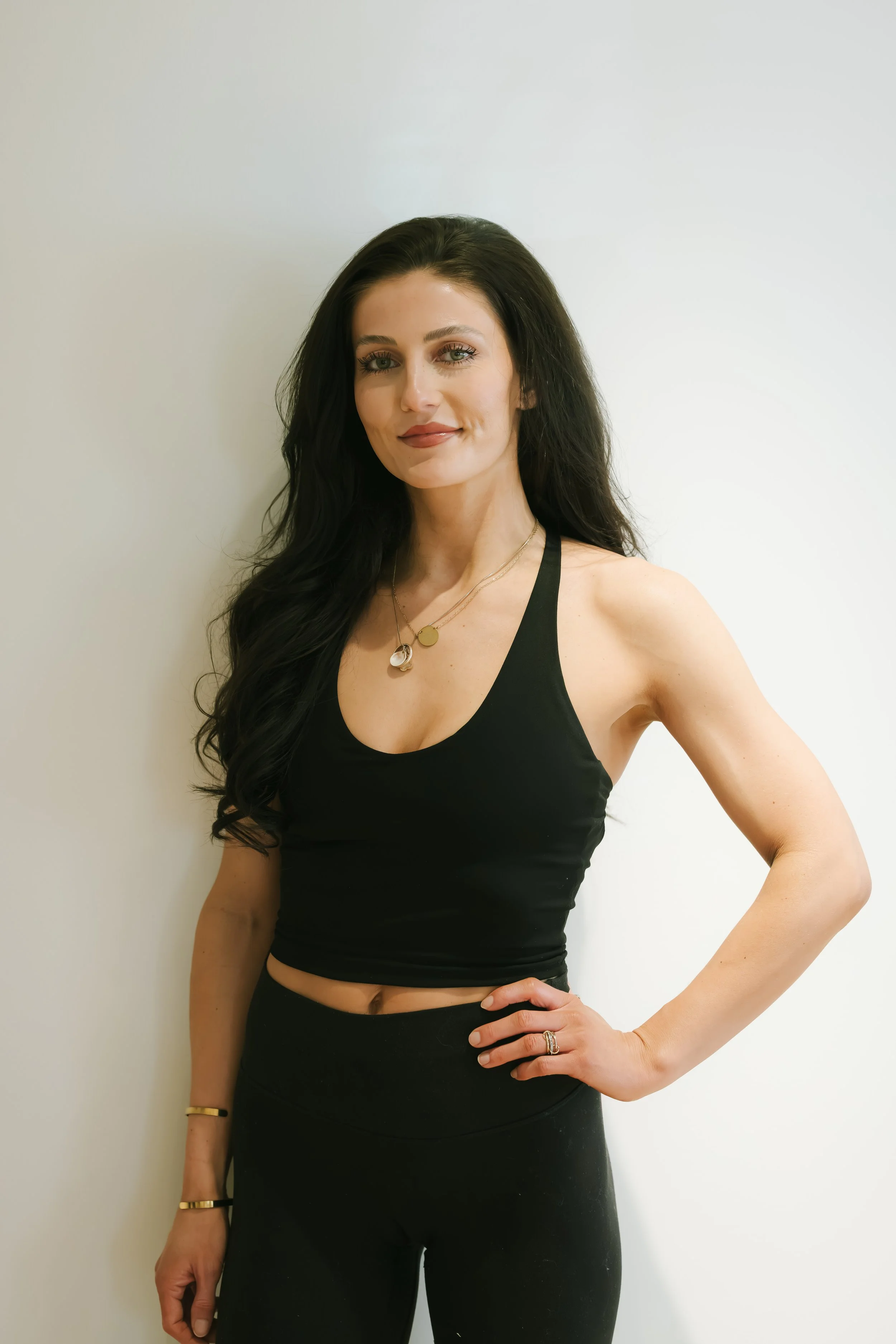 A woman with long dark hair in a black tank top and black pants standing against a white wall, smiling gently, with her right hand on her waist.