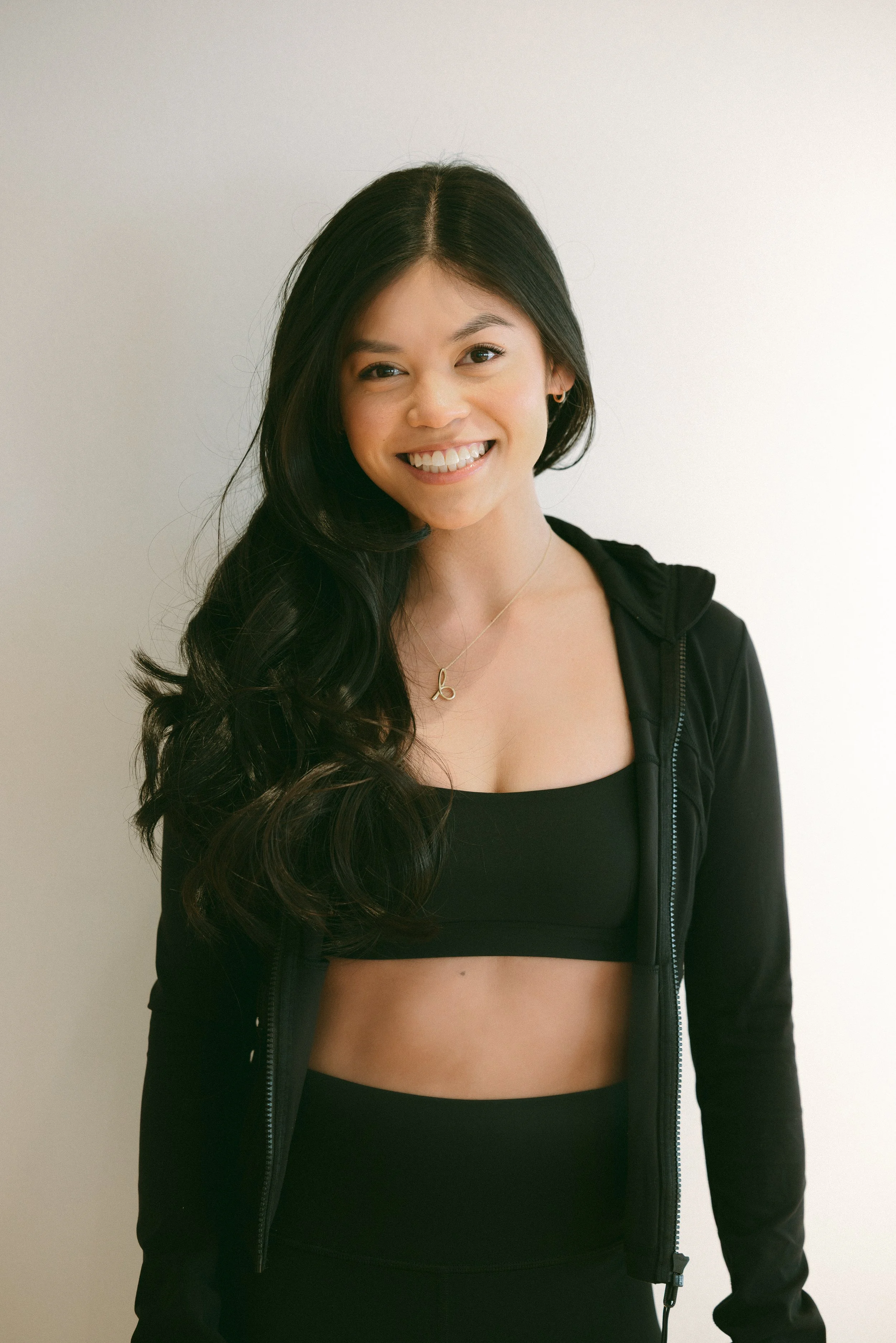 A young woman with long, dark, wavy hair smiling and wearing a black sports bra, black jacket, and a necklace, standing against a plain light-colored wall.