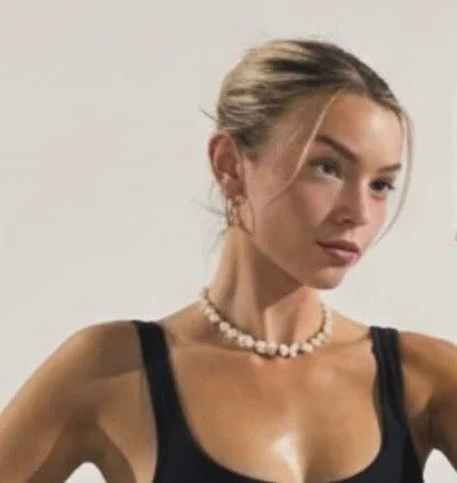 A woman with blonde hair in a bun, wearing a black top and a pearl necklace, posing against a plain white background.