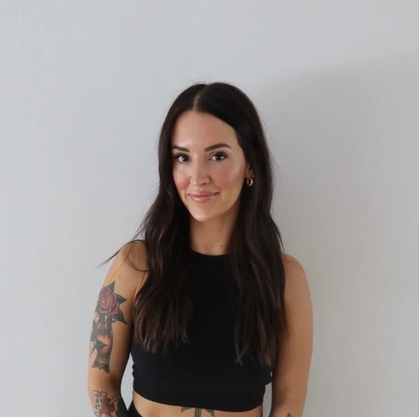 A young woman with long dark wavy hair, wearing a black sleeveless top, standing against a white wall, smiling slightly, with tattoos on her left arm.