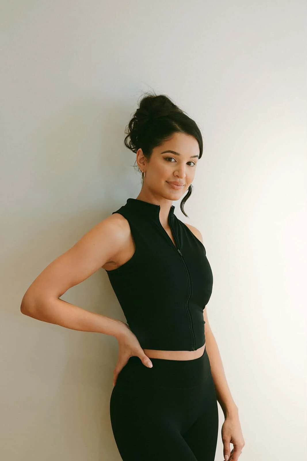 A young woman with dark hair styled in a messy updo, wearing hoop earrings and a black sleeveless zip-up top, standing against a plain white wall and smiling at the camera.