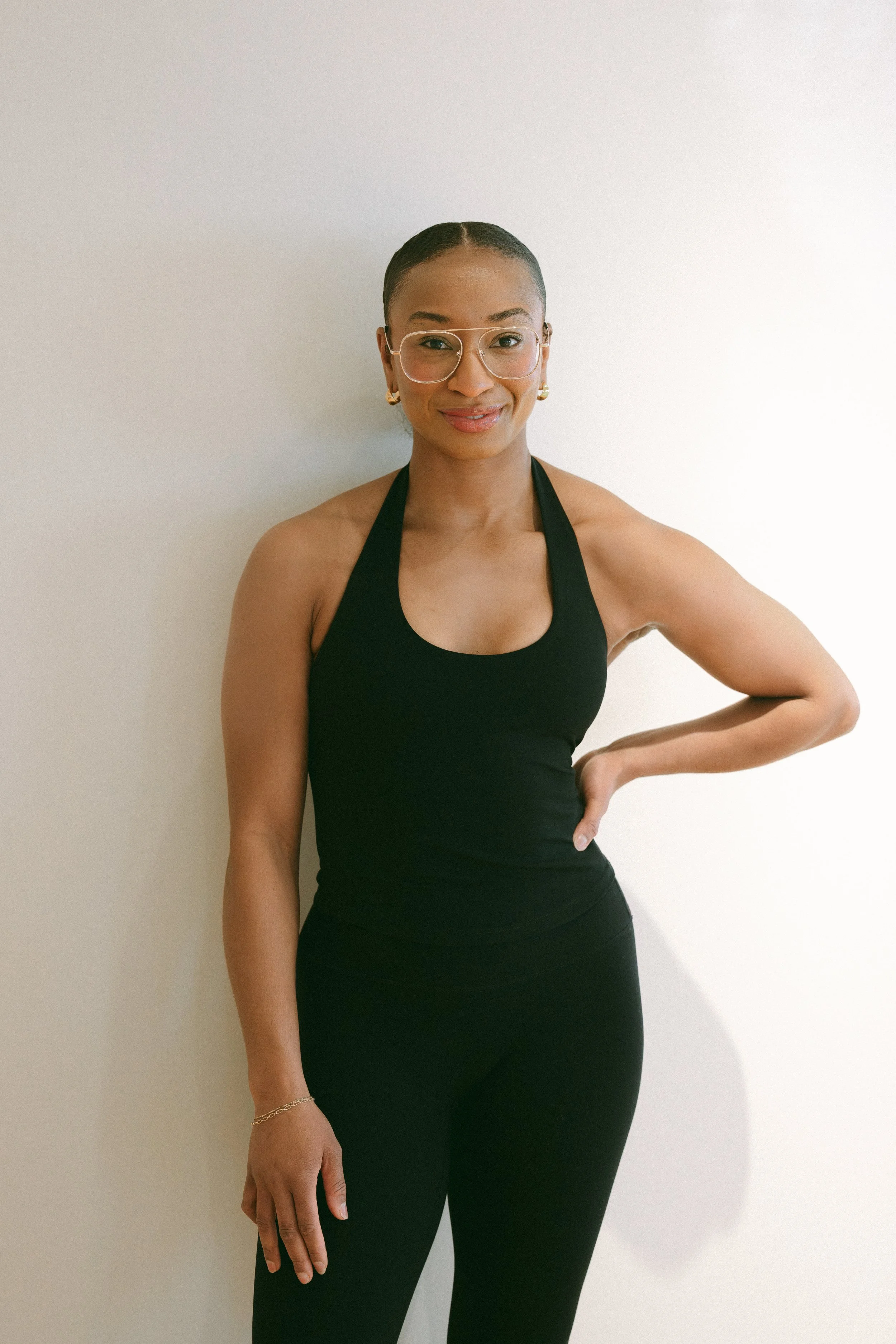 A woman wearing glasses and a black athletic top, standing against a plain white wall, smiling with one hand on her hip.