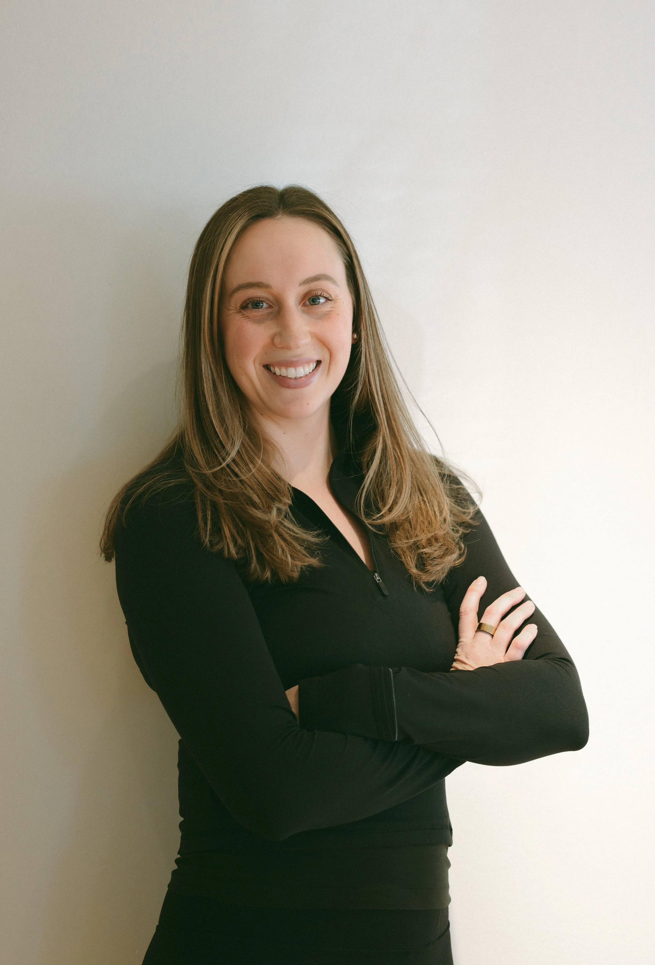 A smiling woman with long light brown hair, wearing a black zip-up top, standing against a plain light background with arms crossed.