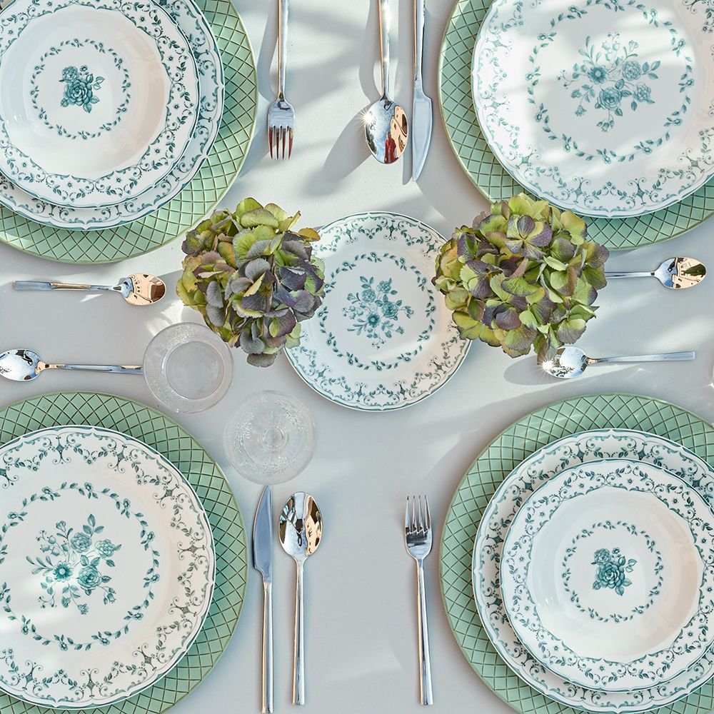 Mesa decorada con platos elegantes con diseño floral, cubiertos de metal, dos centros de flores con hojas verdes, y copas de cristal.