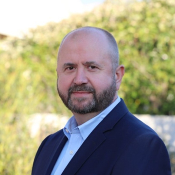 Portrait of a man with a bald head and beard, wearing a navy blazer and light blue shirt, standing outdoors with greenery in the background.