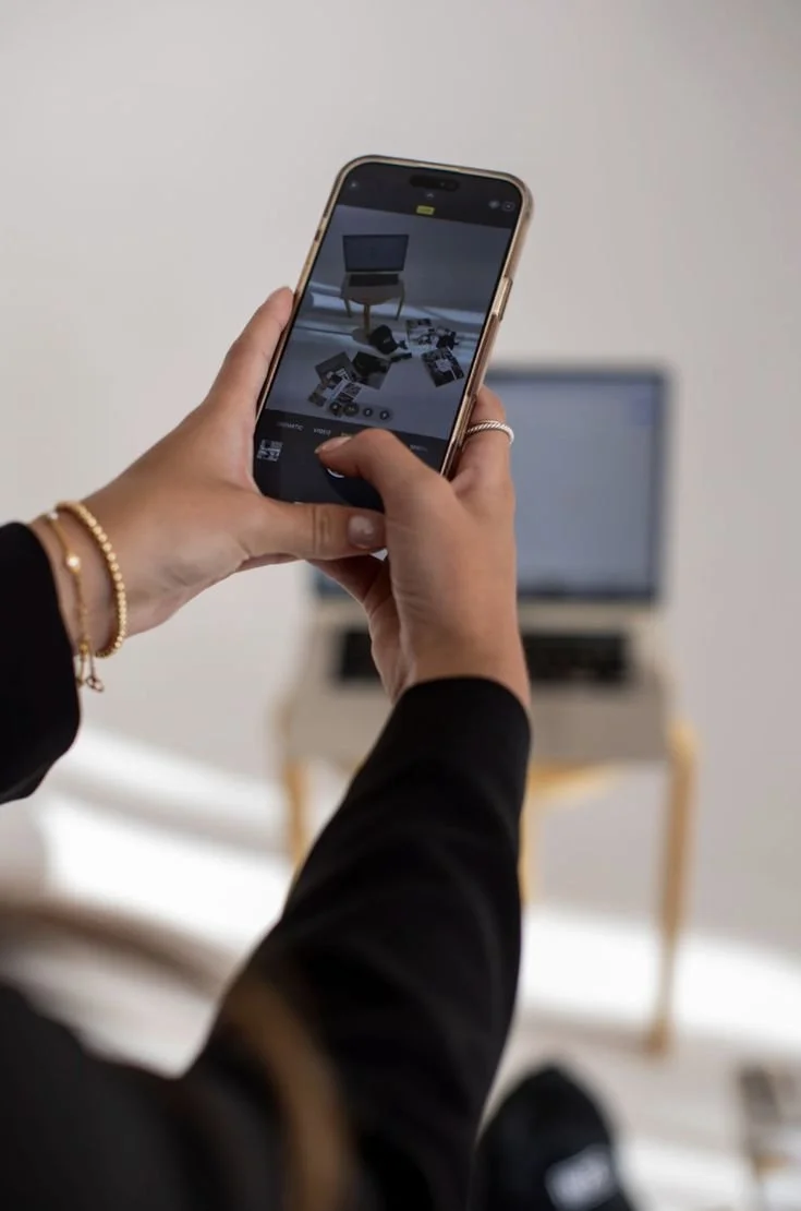 Person taking a photo of a cluttered surface with an iPhone, with a blurred laptop in the background.