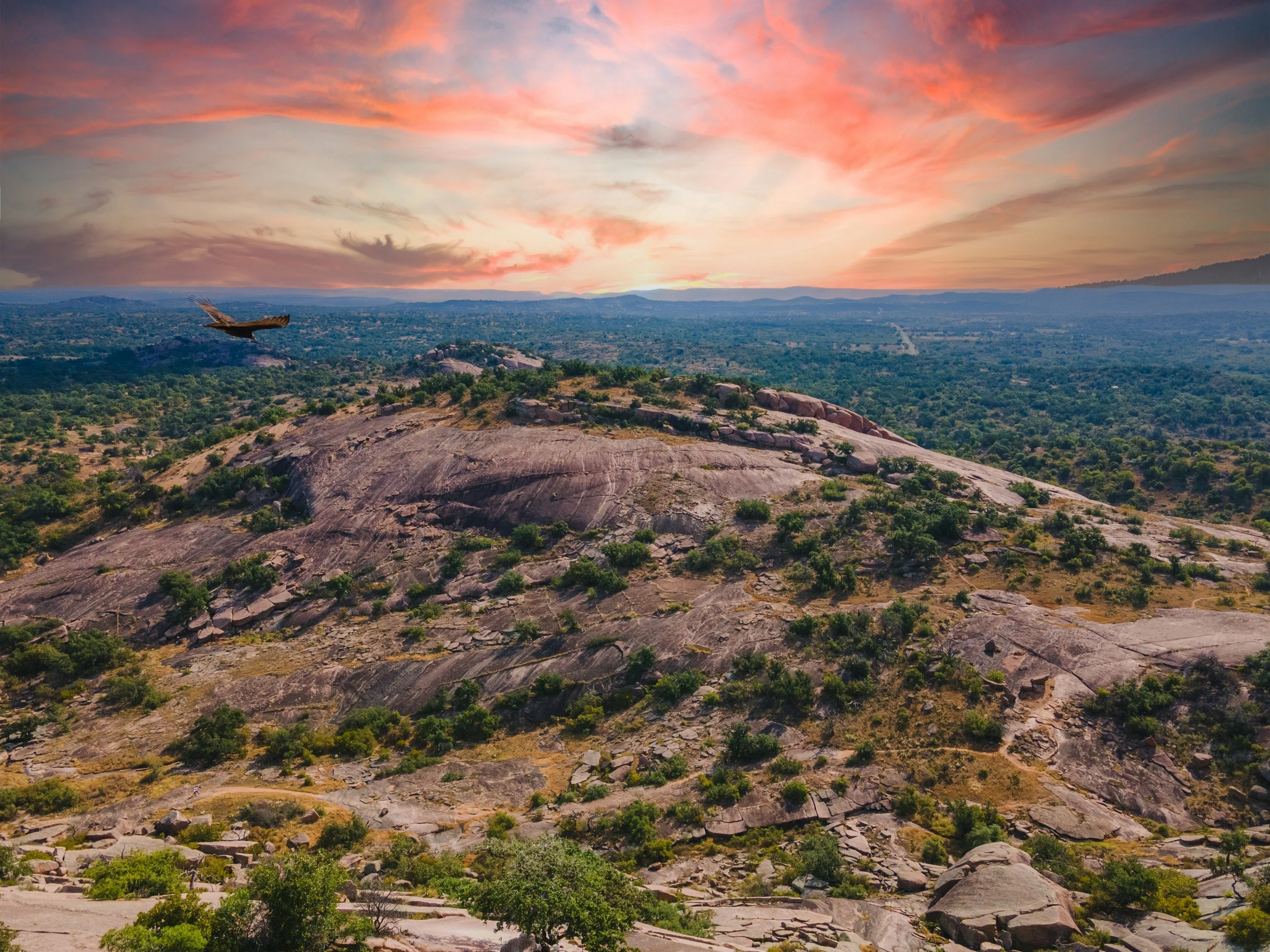 A landscape view of rocky terrain with sparse green vegetation, an eagle soaring in the sky, and a colorful sunset with pink, orange, and purple hues.