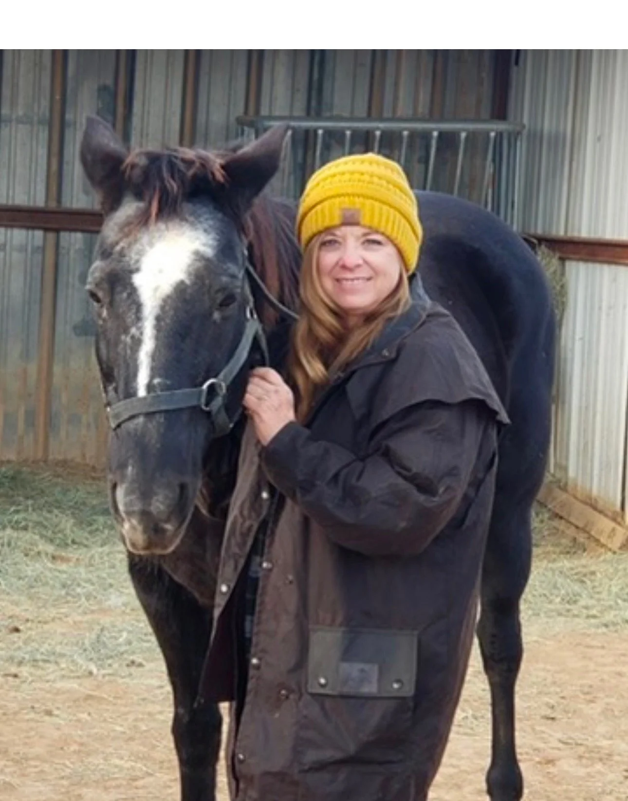 A woman with long hair, wearing a yellow knit hat and a dark brown coat, standing next to a black and white horse inside a barn.