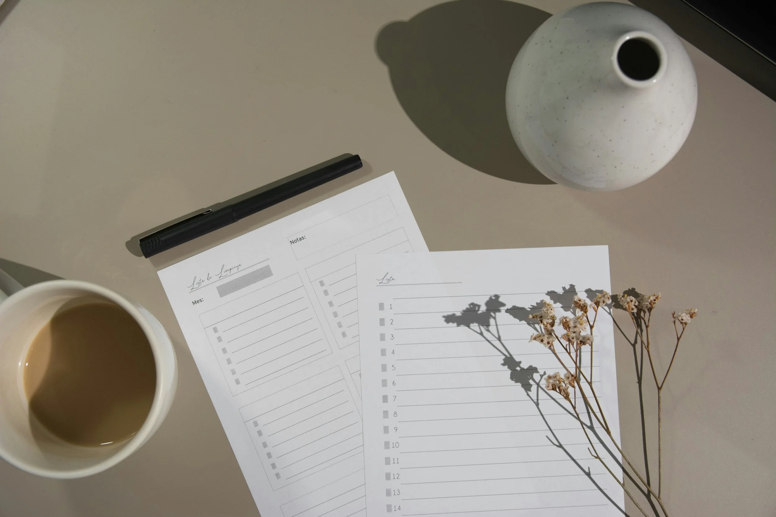 Top-down view of a desk with a black pen, a cup of coffee with milk, two sheets of paper with checklists and writing spaces, a white round vase with a narrow opening, and dried flowers casting shadows on the paper, all against a beige surface.