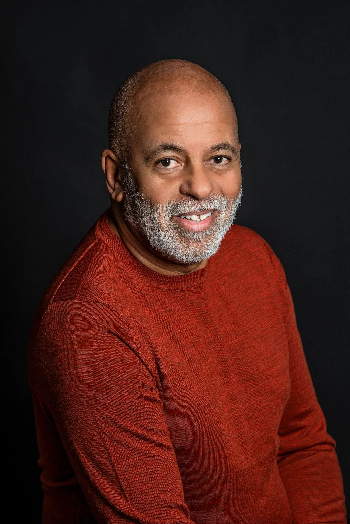 Portrait of a smiling middle-aged African American man with gray beard, wearing a red sweater, against a black background.