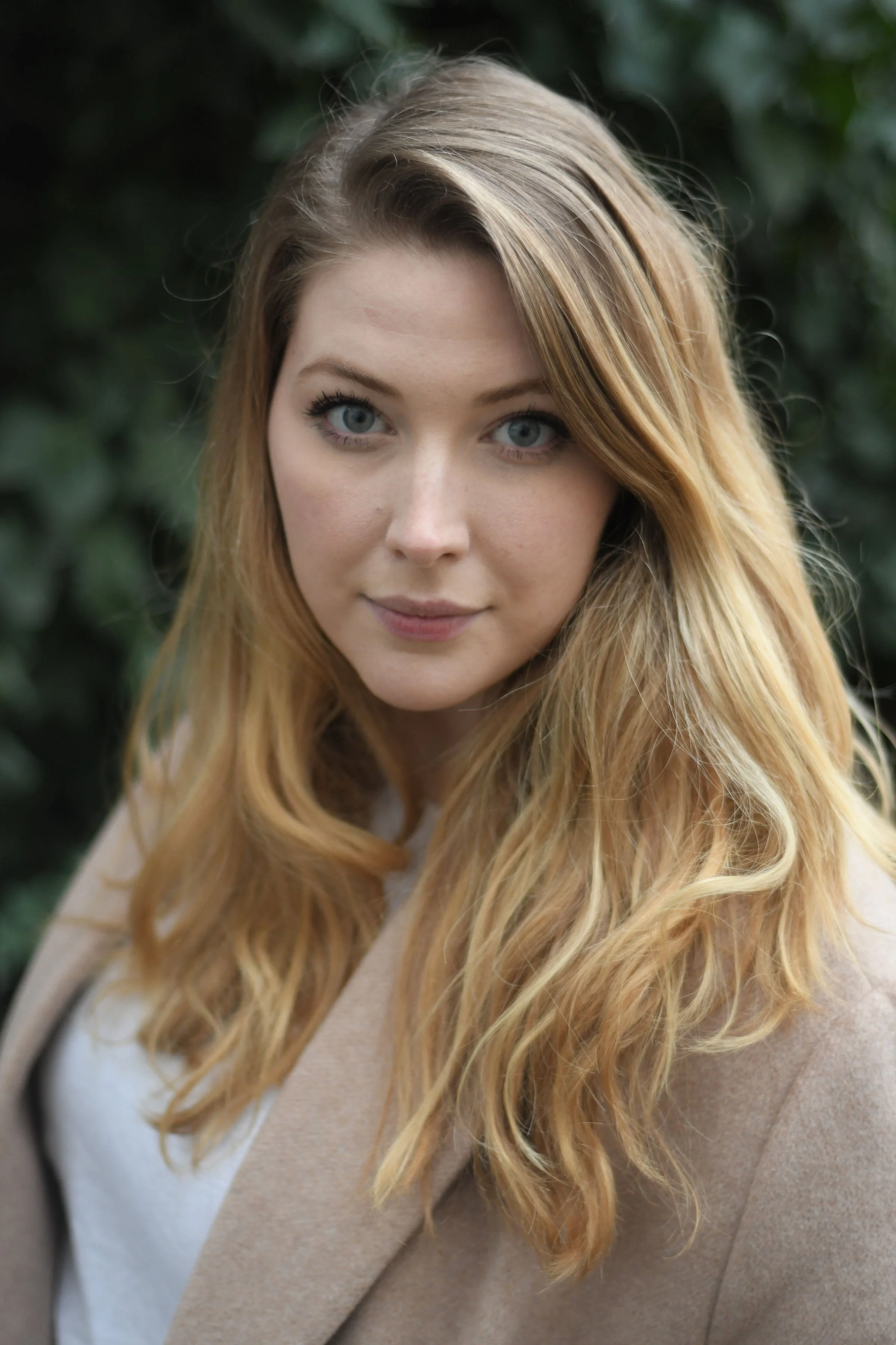 Close-up of a woman with long, wavy blonde hair, blue eyes, and light skin, outdoors with green foliage in the background.