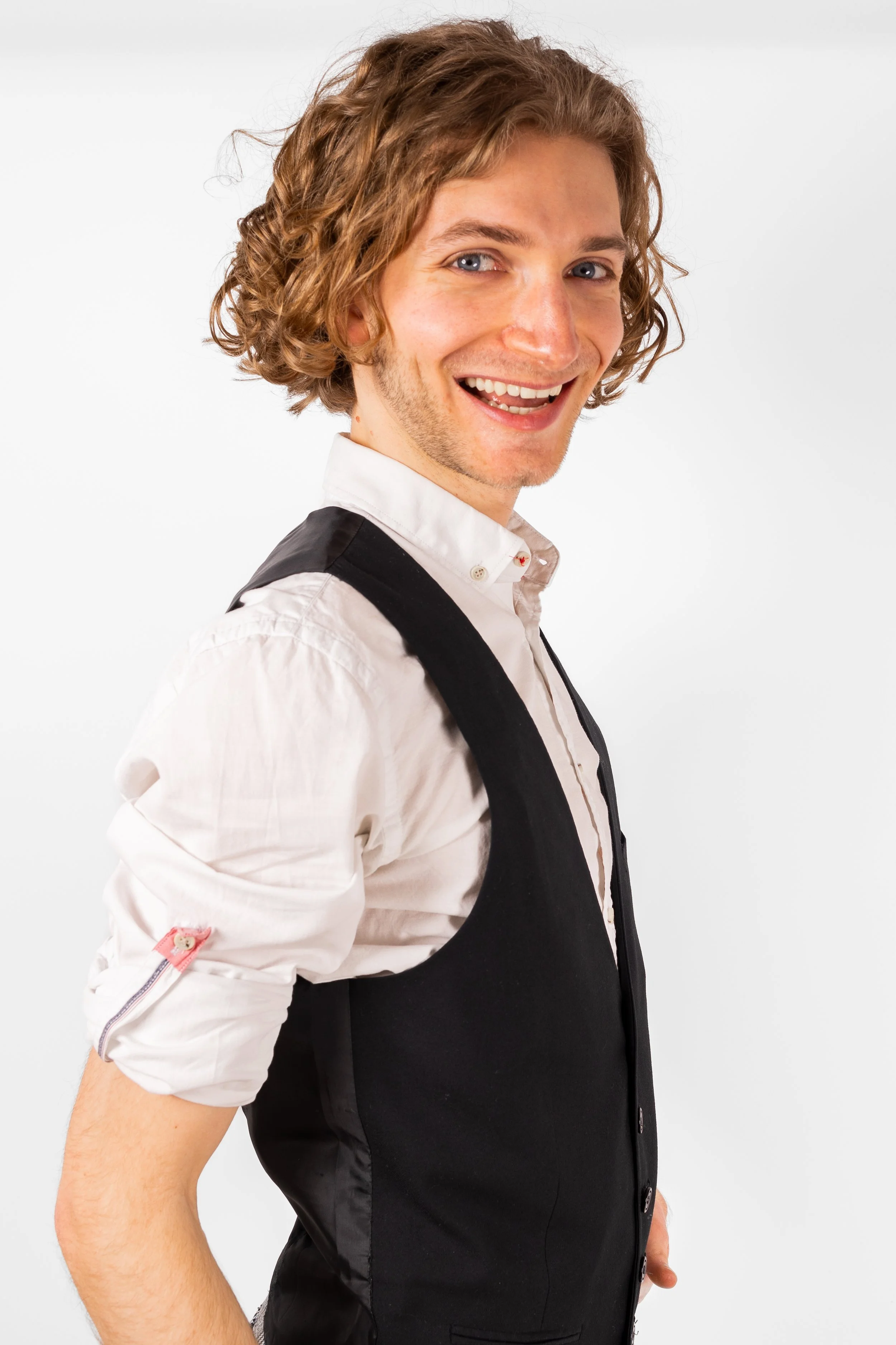 A young man with curly blond hair smiling in a professional studio setting, wearing a white dress shirt and a black vest.
