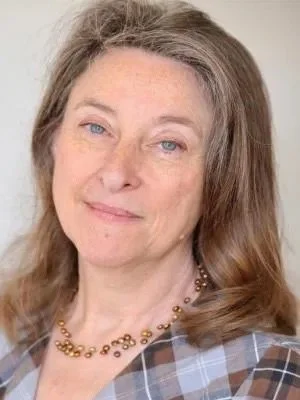 A portrait of a woman with light brown hair and blue eyes, wearing a plaid shirt and a beaded necklace.