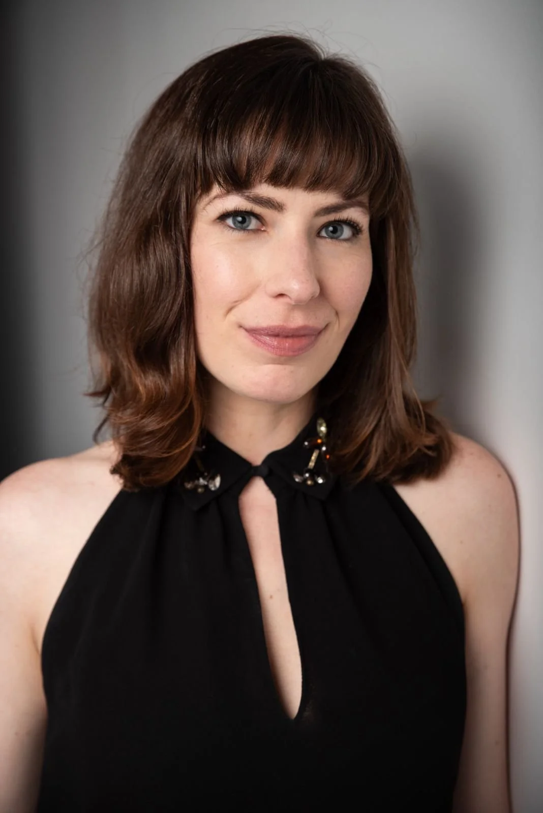 A woman with shoulder-length wavy brown hair and blue eyes wears a sleeveless black dress with a collar decorated with small embellishments and a keyhole neckline. She smiles softly against a neutral background.