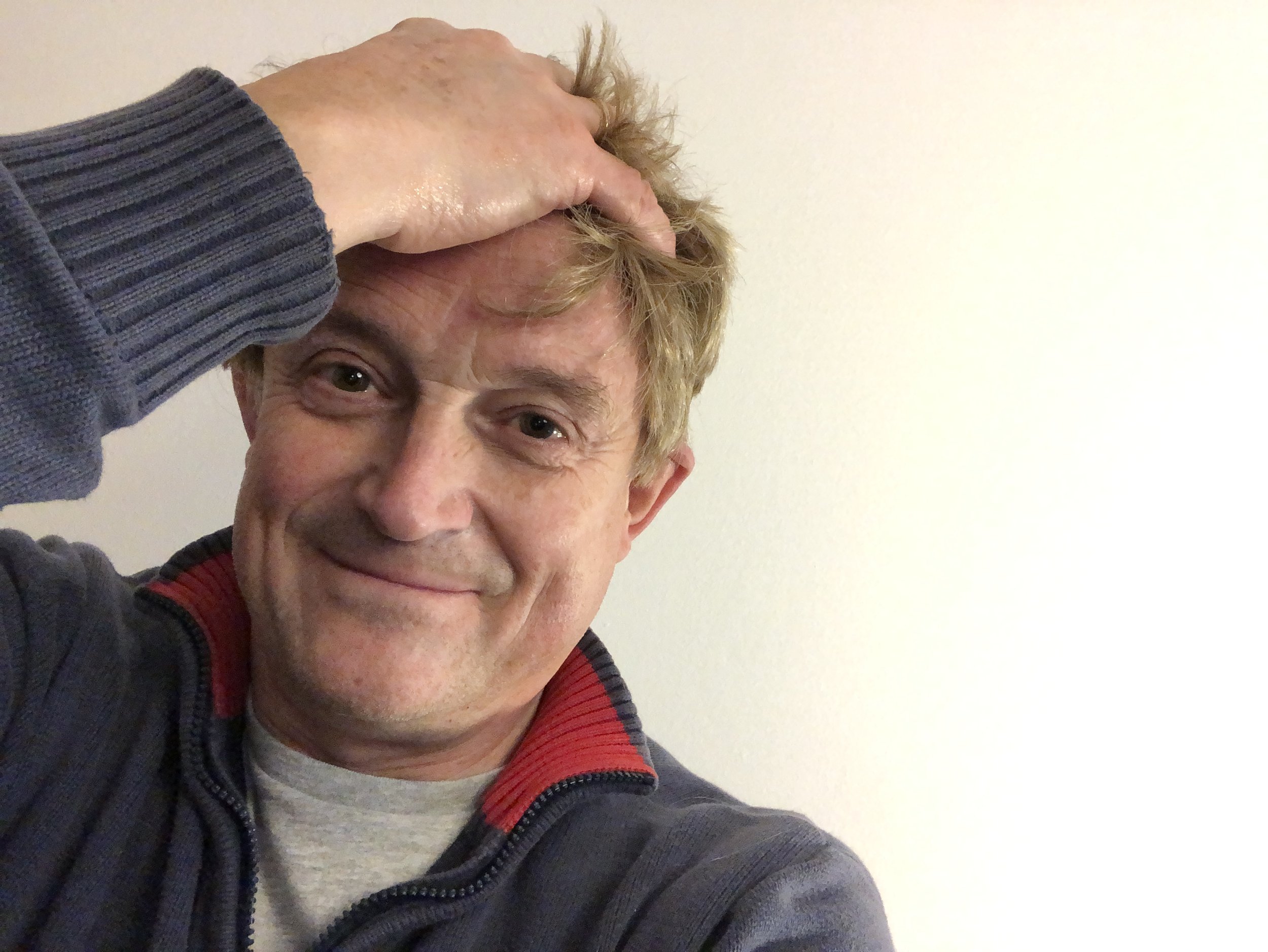 A man with light-colored, tousled hair is touching his forehead with his right hand. He is smiling and wearing a dark-colored jacket with a gray shirt underneath.