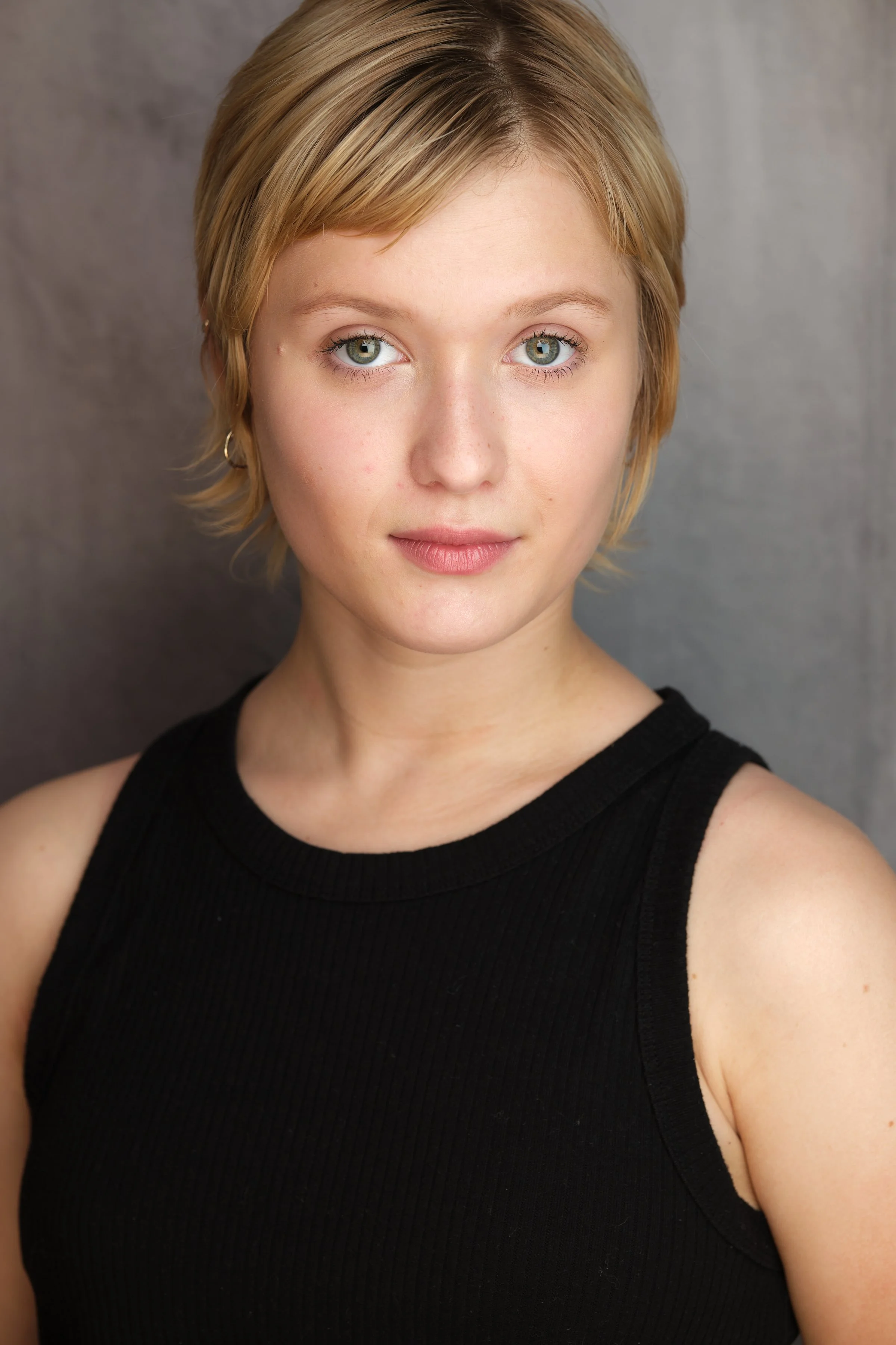 Close-up of a young woman with short blonde hair and light blue eyes, wearing a sleeveless black top, standing against a gray background.