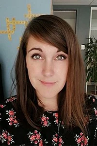 Portrait of a woman with straight brown hair and bangs, wearing a black floral top, standing indoors with a light blue wall and a plant in the background.