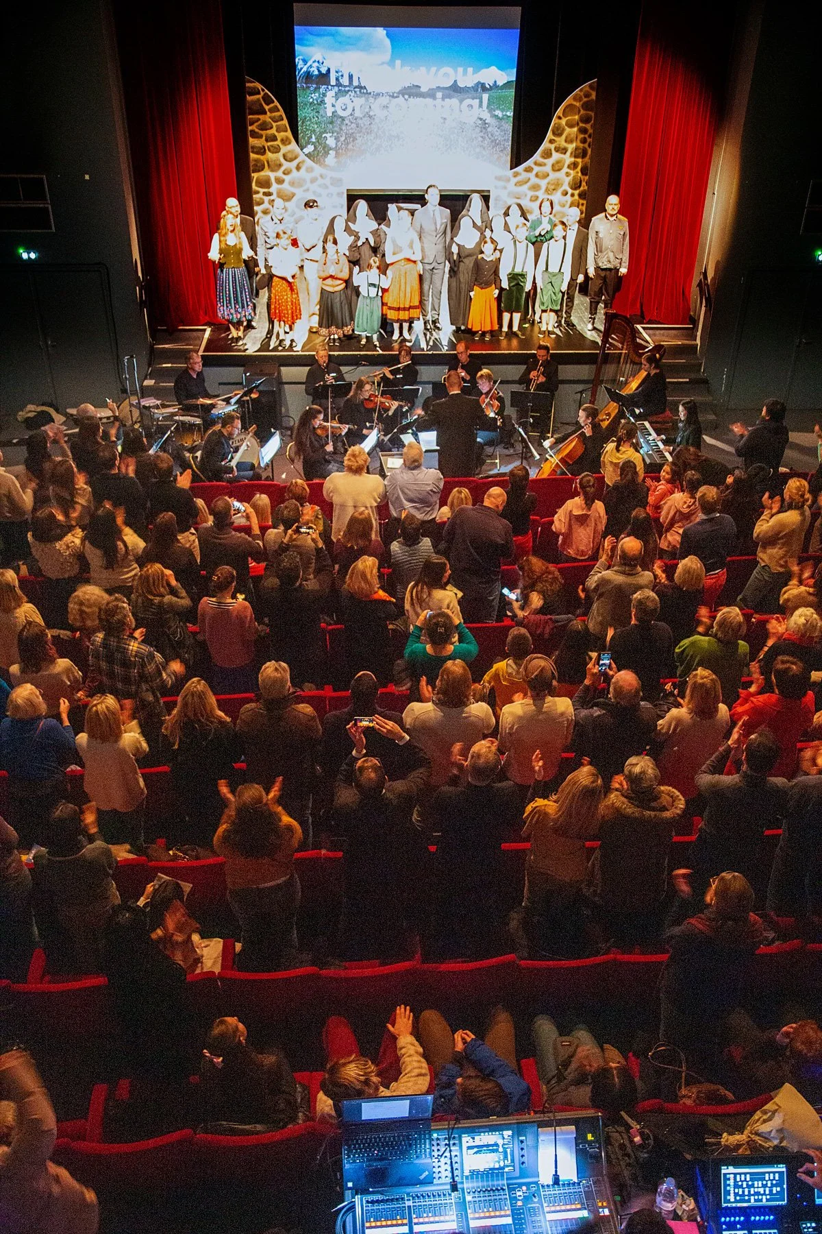 People watching a theater performance on stage with a group of actors and musicians in front of a large screen.