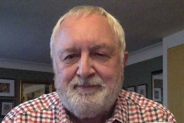 A close-up of a man with gray hair and a beard, wearing a red and white checked shirt, smiling softly inside a room with framed pictures on the walls.