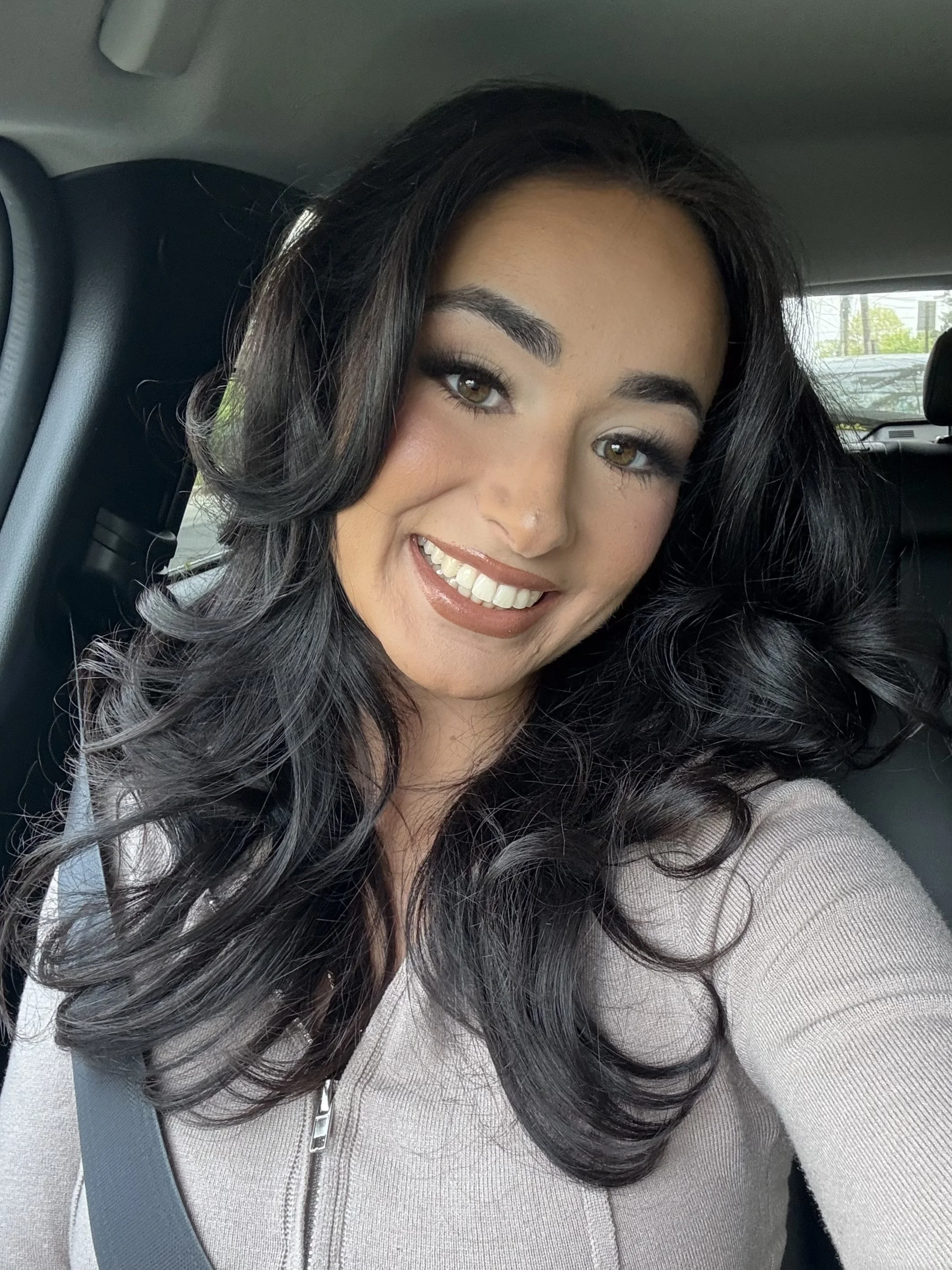 A woman with long, wavy black hair smiling inside a car, with visible seatbelt and a blurred background of parked cars and trees.