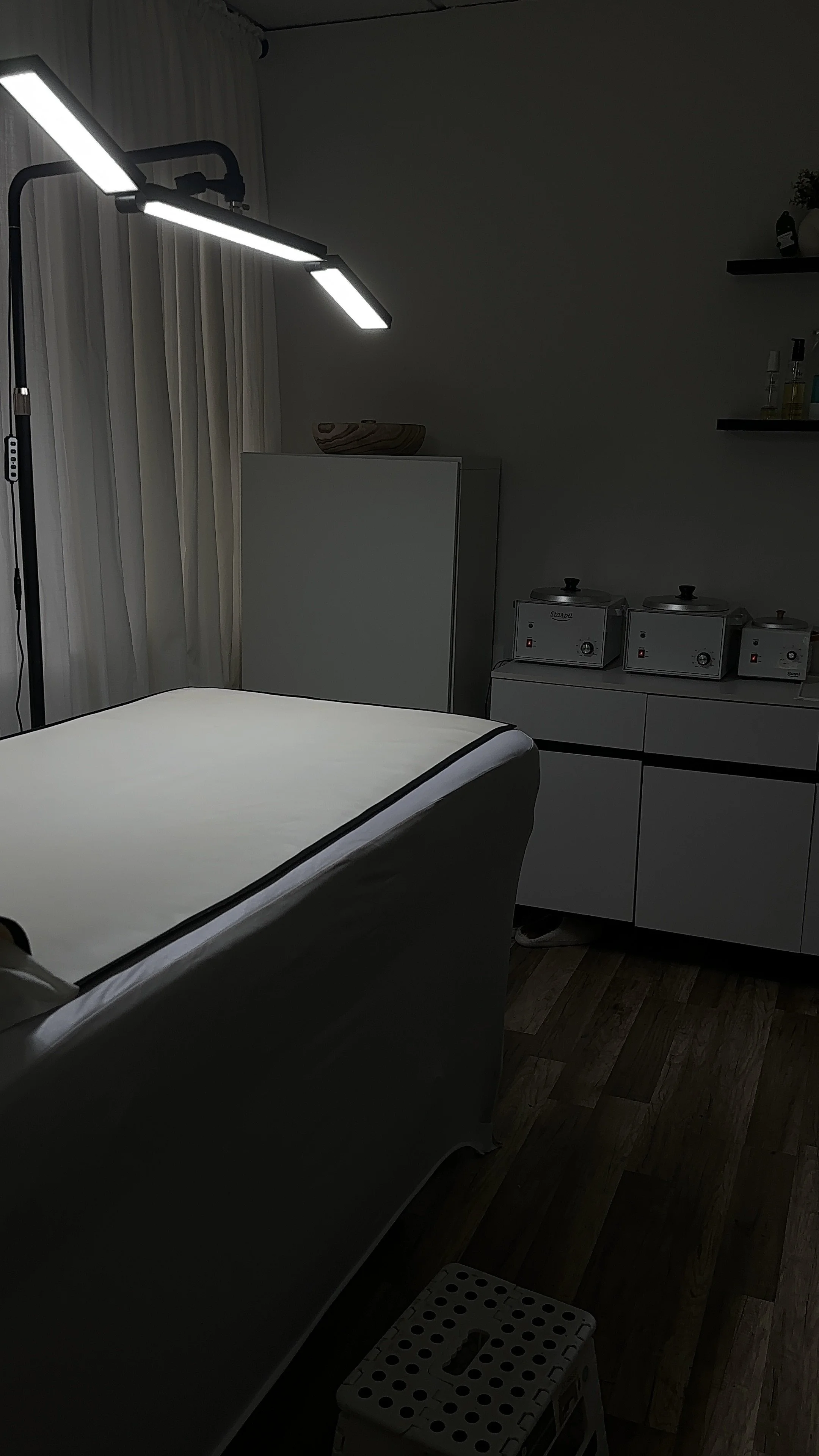 A modern, dimly lit spa or massage room with a massage table covered with a white sheet, a large LED light source overhead, white cabinets, and infrared heaters or steamers on a counter.