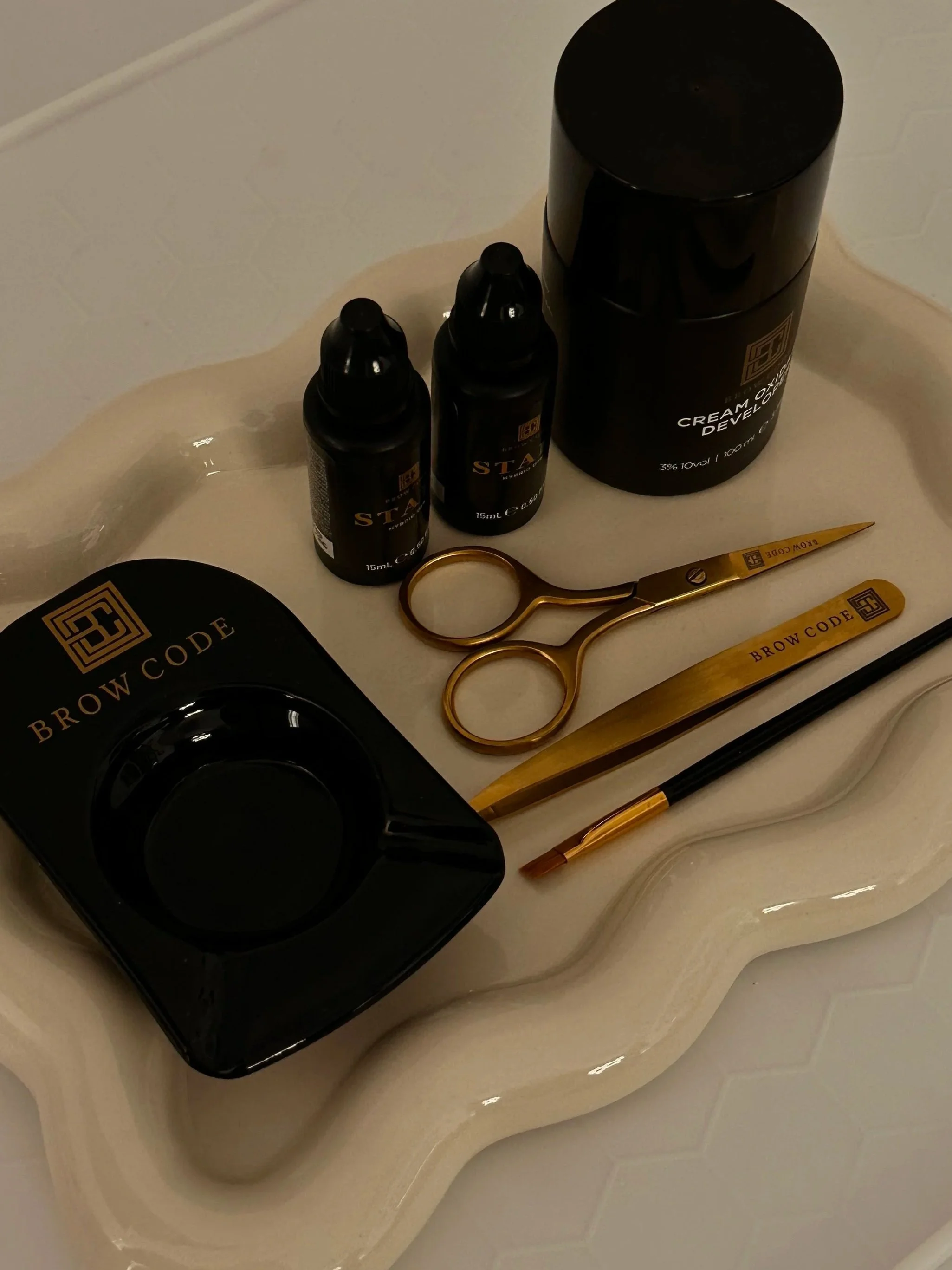 Black tray labeled 'Brow Code' contains tattoo or microblading pigment bottles, scissors, a sowing needle, a pen, and tweezers, all arranged on a cream-colored wavy tray.