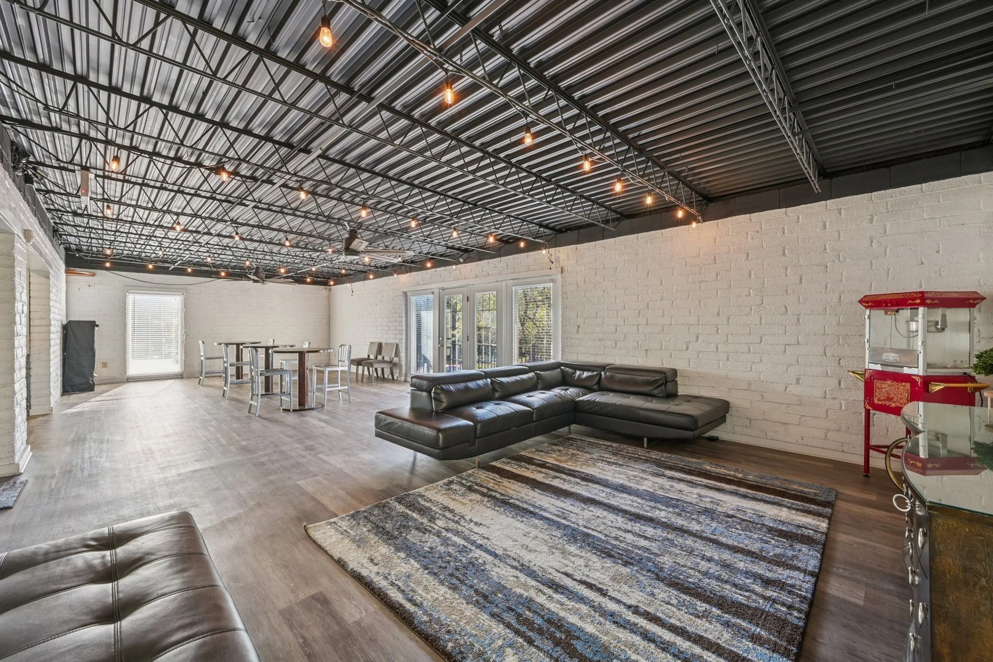 Open living space with black leather sectional sofa, white brick walls, wooden flooring, string lights hanging from black metal ceiling, and a rug with abstract blue, gray, and cream pattern. Furniture includes a bar-height table with chairs, a popcorn machine, and a glass-top side table.