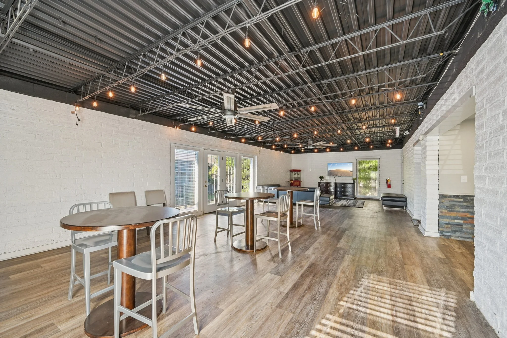 Spacious indoor area with white brick walls, wood flooring, string lights on a black metal ceiling, and a ceiling fan. There are tables and chairs, a TV, a couch, and a small kitchen area with a popcorn machine.