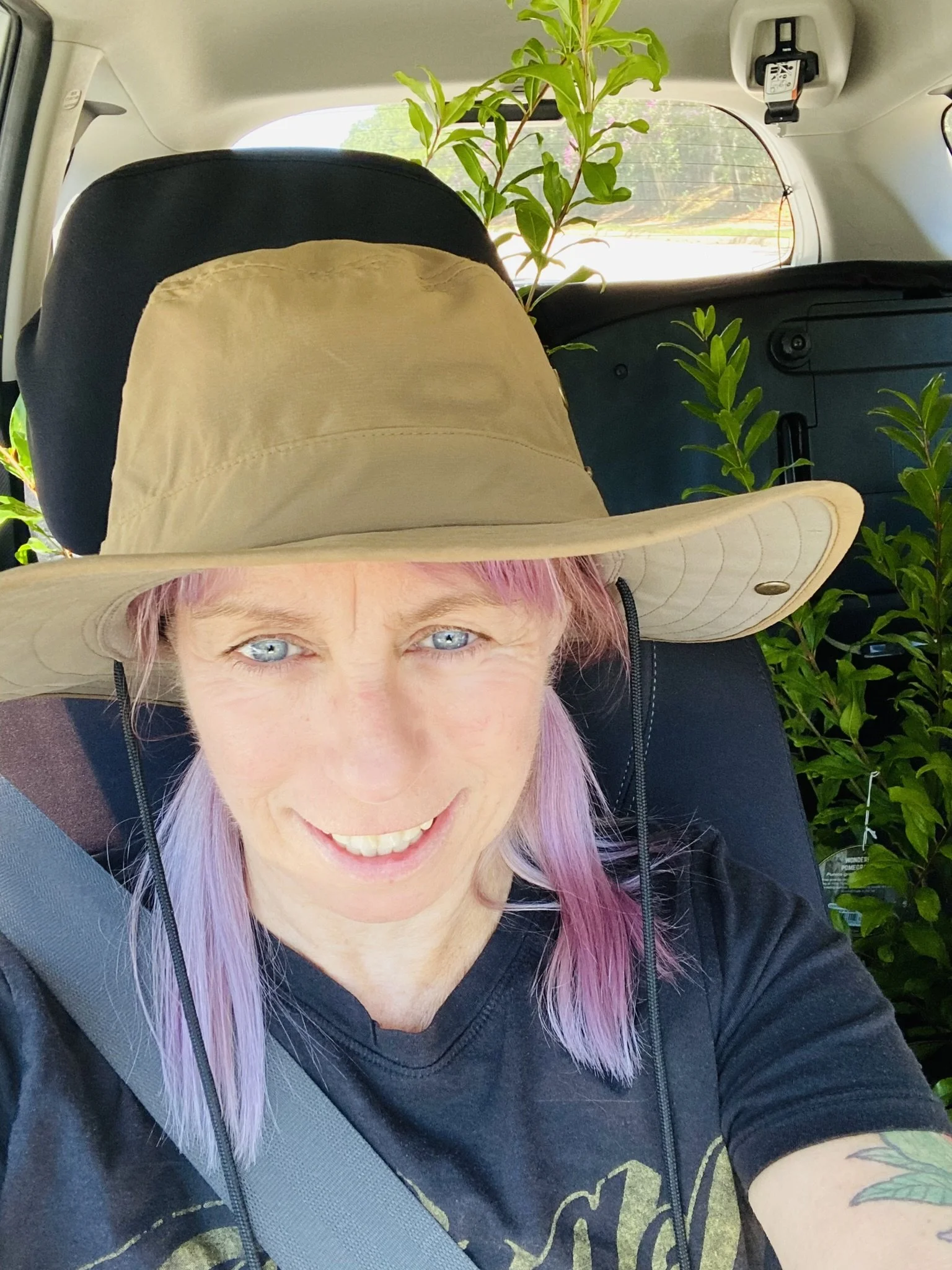 A woman with purple hair, blue eyes, and a tattoo on her arm smiling inside a vehicle. She is wearing a wide-brimmed hat and a black T-shirt, with plants and greenery visible in the background.