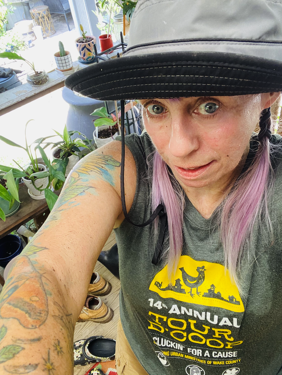 A woman with purple hair and tattoos is taking a selfie, wearing a cap and a T-shirt with the logo for the 14th Annual Tour d'Coop event, in an indoor space filled with potted plants and natural light.