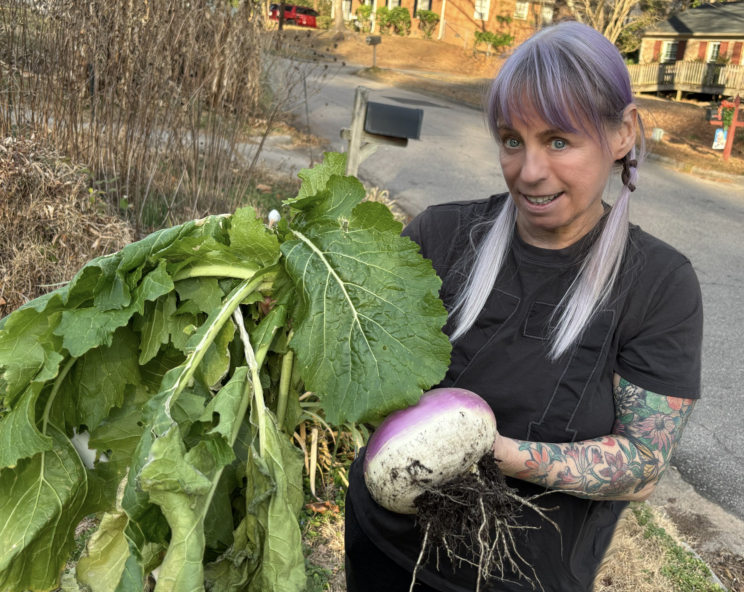 A woman with purple hair and tattoos on her arm is holding a large, freshly harvested turnip with dirt on its roots. She is standing outdoors next to a leafy green plant and smiling at the camera, with a street and houses in the background.