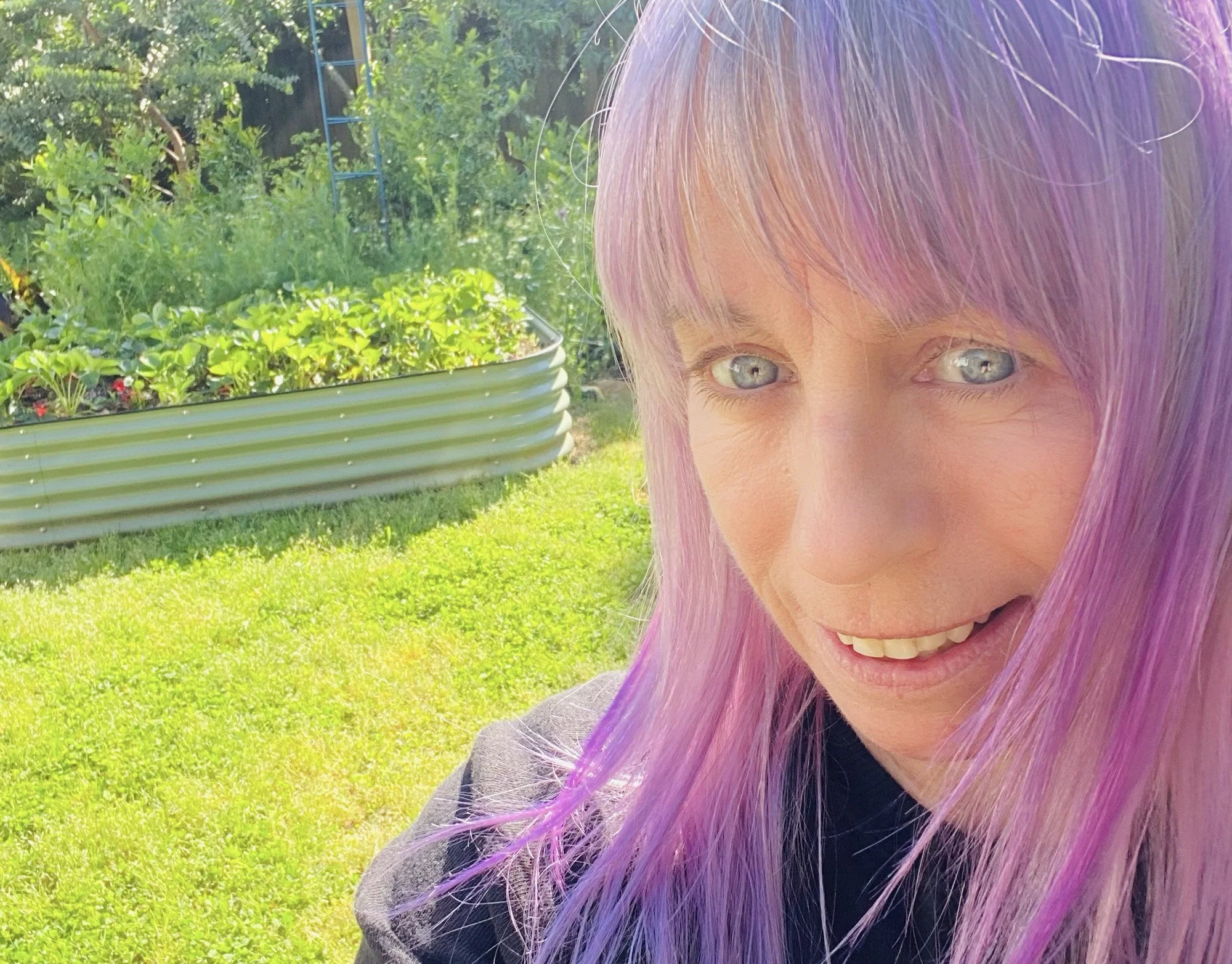Ralstead Foodscapes founder, Ingrid, smiling in a garden with green grass and a raised garden bed filled with plants in the background.