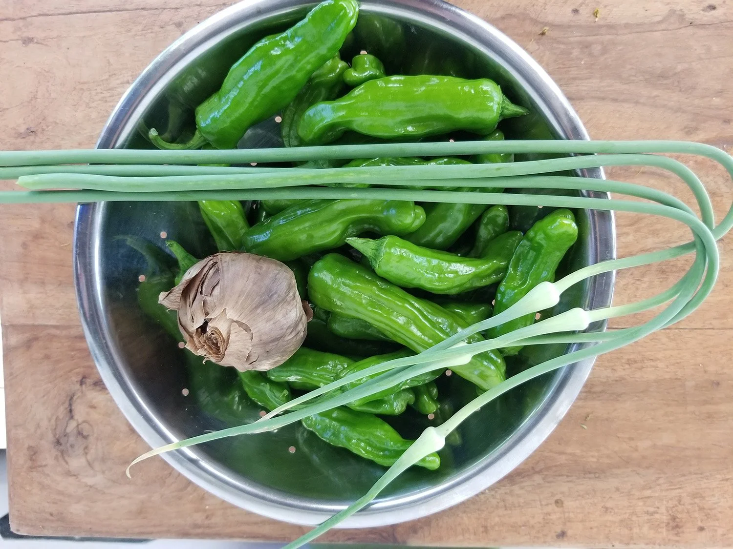 A metal colander filled with green chilies, a garlic bulb, and green onions on a wooden surface.