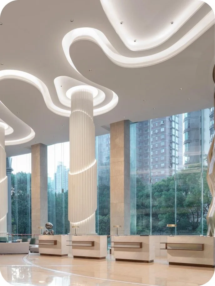 Luxurious hotel lobby with tall, white, spiral columns, modern ceiling design with ambient lighting, large glass windows showcasing city buildings and green trees outside.