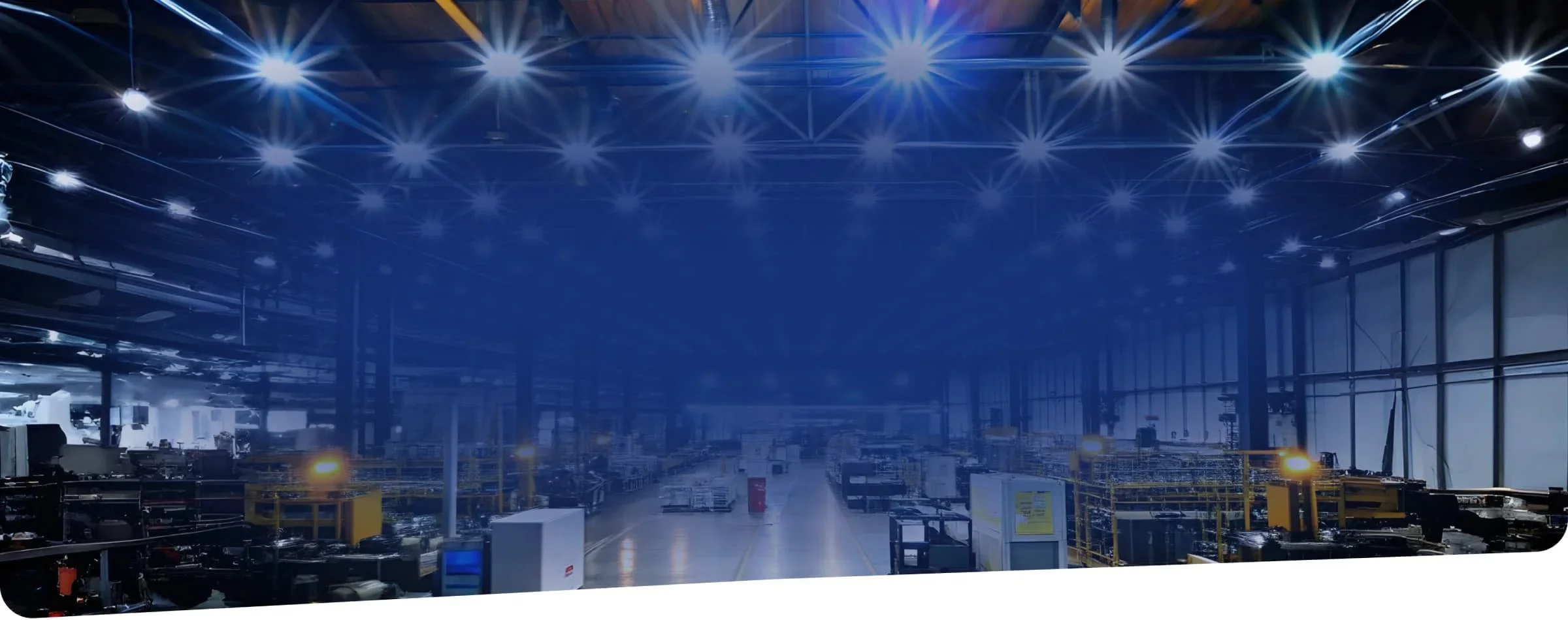View of a large, empty industrial warehouse with high ceilings, blue lighting, and various equipment and conveyor belts.
