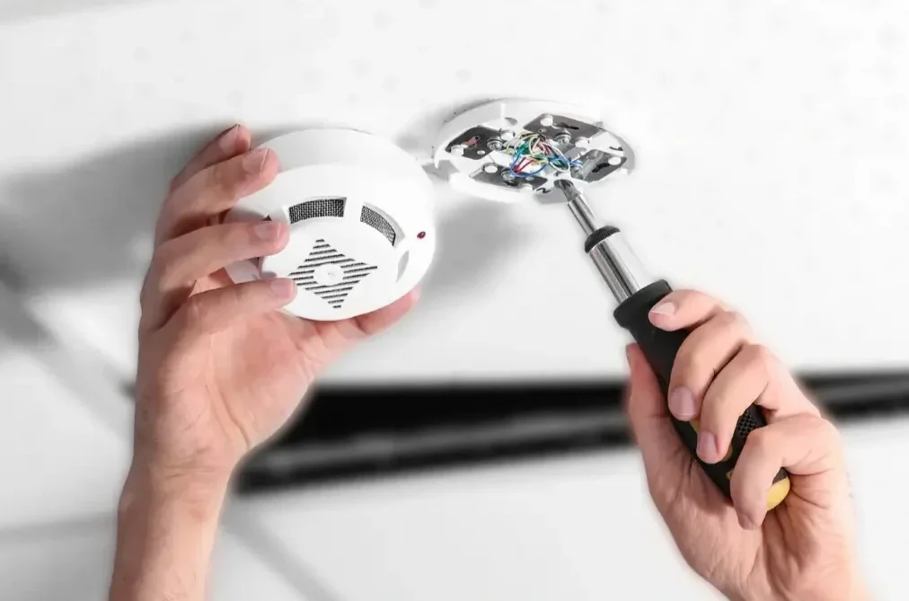 A person installing or repairing a smoke detector on a ceiling with a screwdriver.