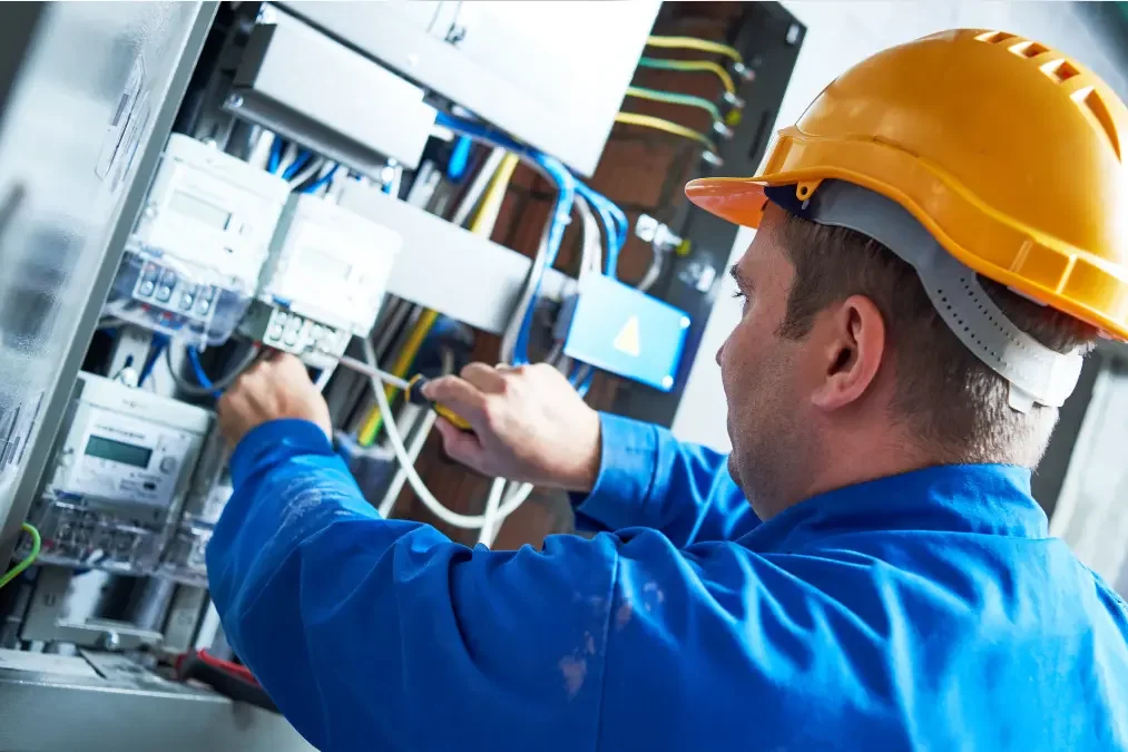 A male electrician wearing a yellow safety helmet and blue work shirt working on electrical wiring inside an electrical panel or control box.
