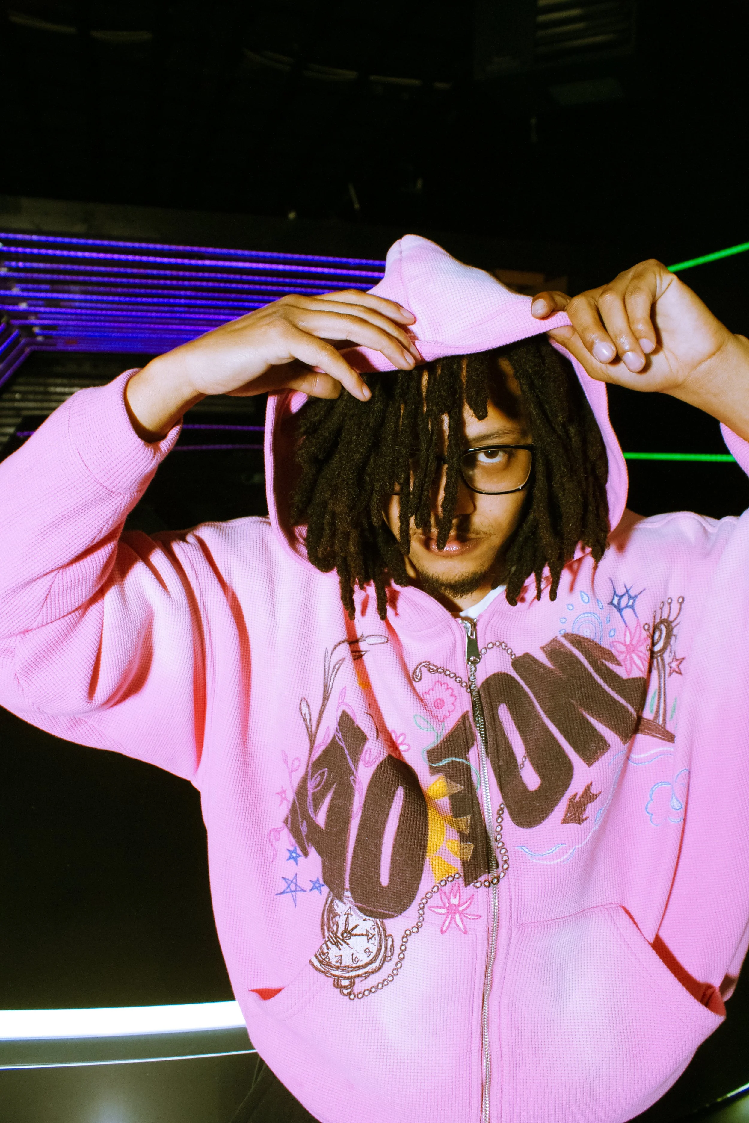Person with dreadlocks wearing glasses in a pink hoodie, holding the hoodie with both hands, standing indoors with colored lights in the background.