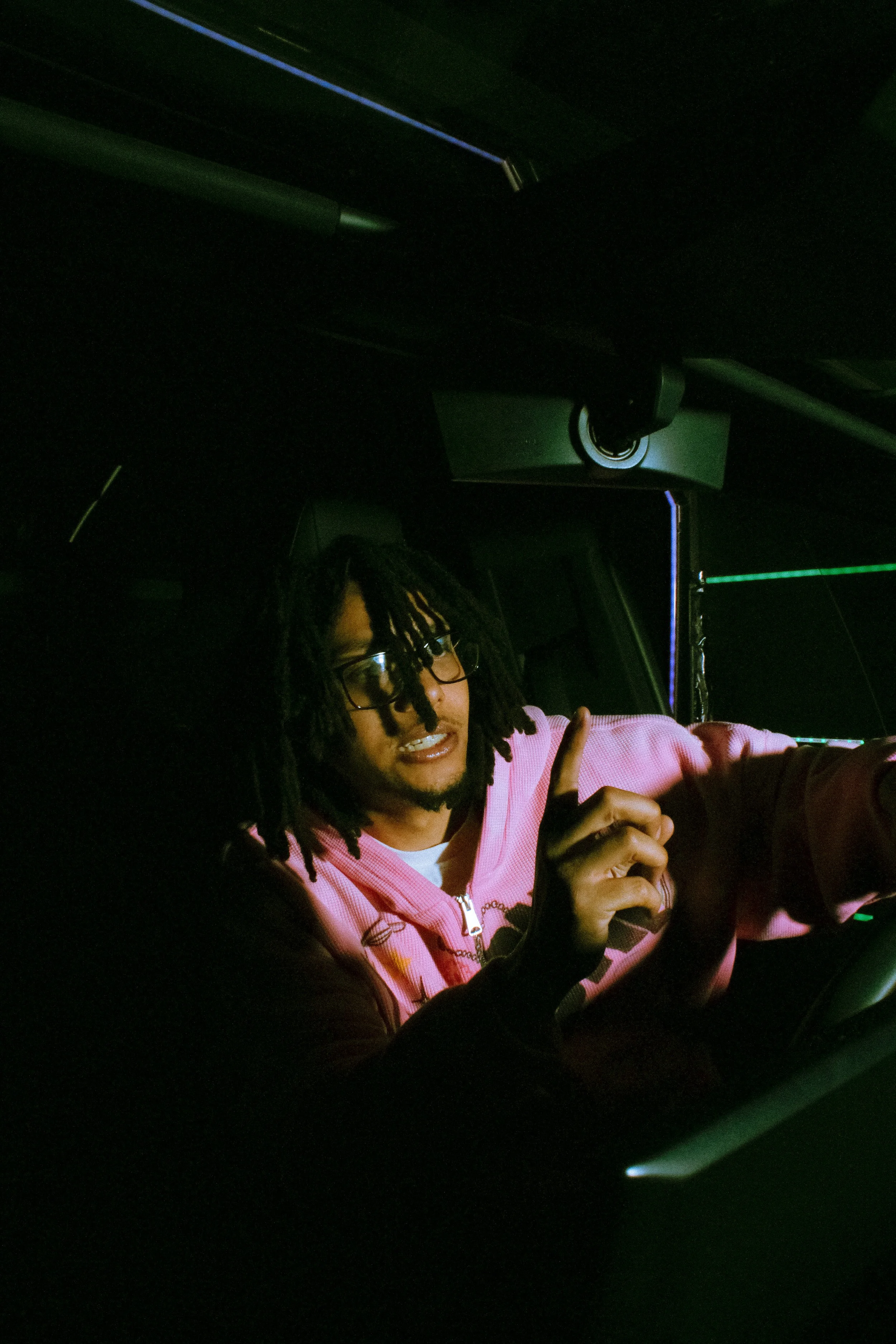 A person with glasses and dreadlocks wearing a pink hoodie, smiling and making a peace sign with their hand inside a dark car.