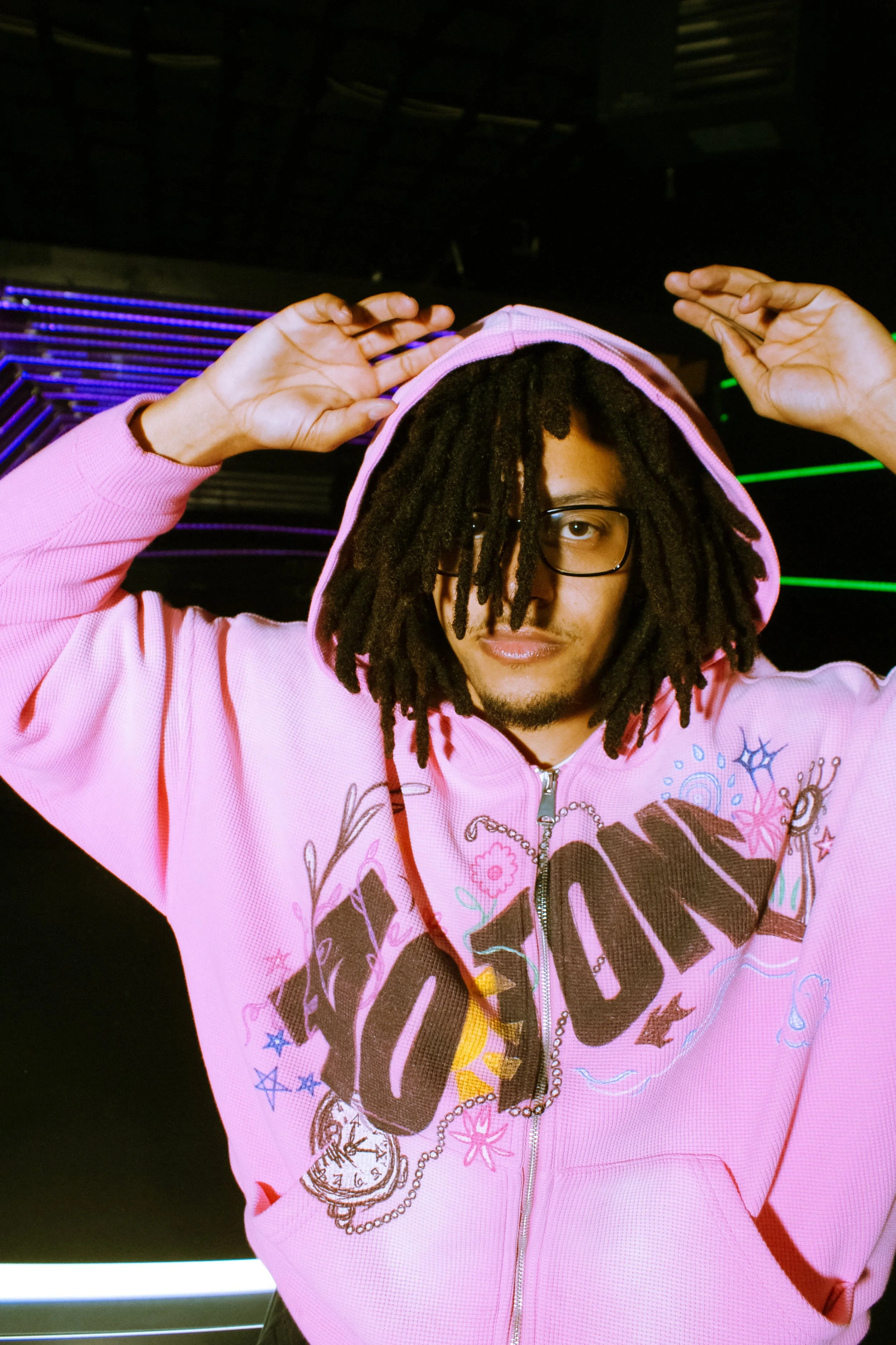 A person with dreadlocks wearing glasses and a pink hoodie with a graphic design, standing in front of a dark background with purple and green neon lights.